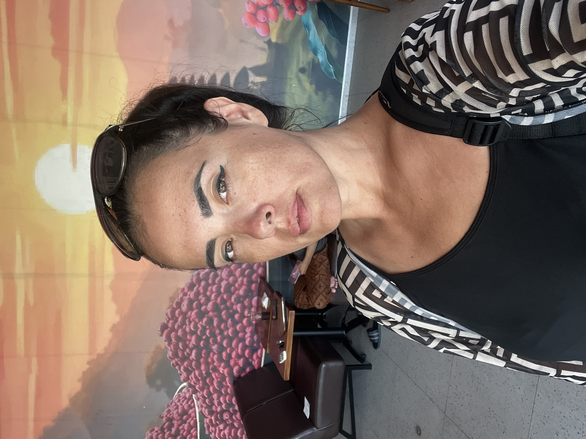 In the city of Kuta Selatan, Indonesia, a woman with dark hair pulled back and sunglasses perched on her head takes a selfie. She is wearing a black tank top and a black and white patterned mesh cardigan. Behind her, a wall mural depicts a vibrant landscape with a large, setting sun, distant mountains, and piles of red berries. To her left, a dark brown booth seat is visible, along with a small table holding a phone, a drink, and a small stack of papers. In the background, another person is partially visible seated on a couch, looking at a phone. The floor is tiled with a gray and white pattern.