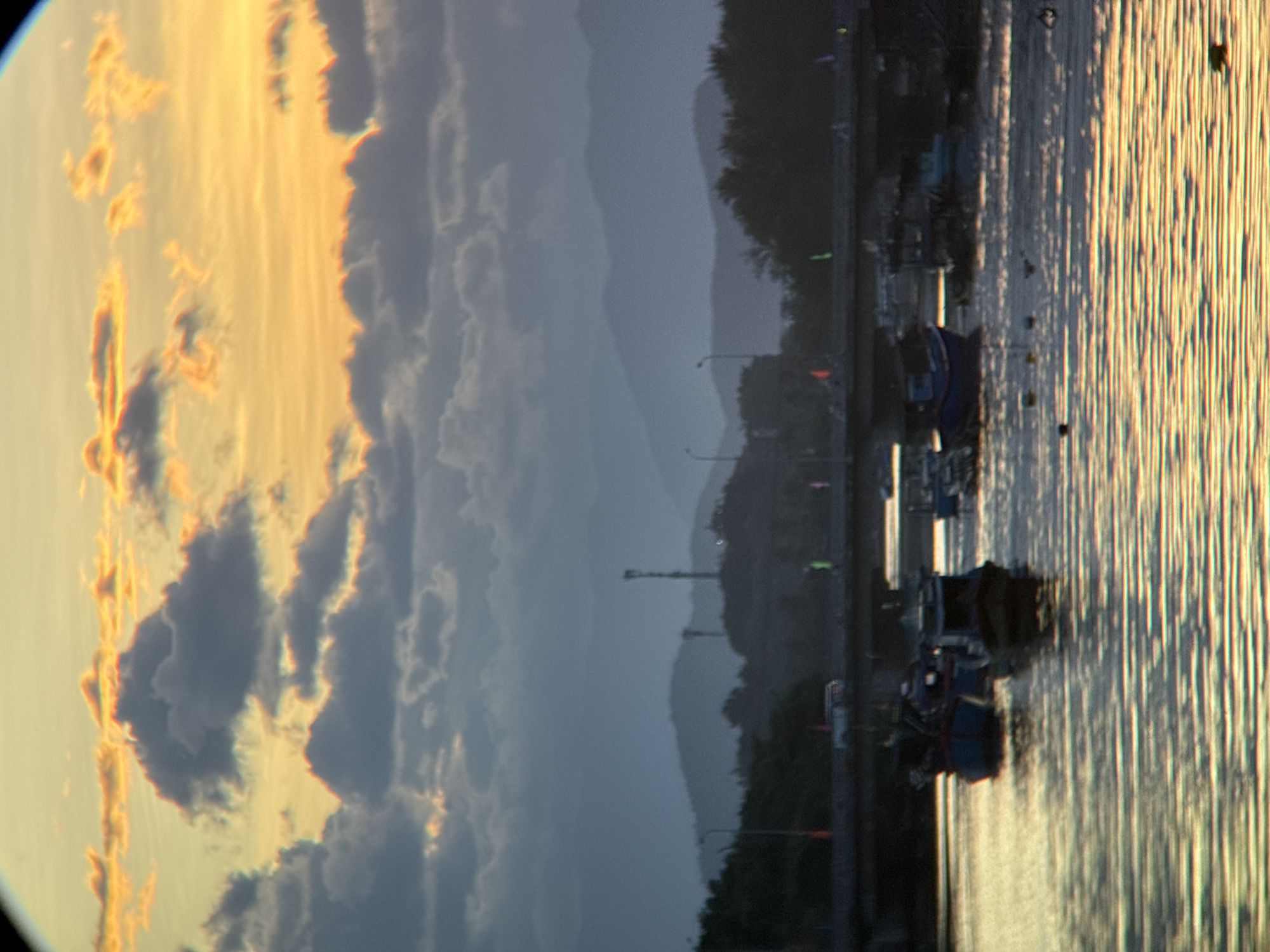 This is a photograph of a scenic landscape near Nha Trang, Vietnam, likely taken during sunset or sunrise. The image captures a body of water, possibly a river or a bay, with several small boats anchored or floating on its surface. A bridge spans across the water in the mid-ground, with vehicles visible on it. Beyond the bridge, a series of hills or mountains are silhouetted against the sky, which is filled with dramatic clouds illuminated by the setting or rising sun. The overall impression is one of tranquility and natural beauty, with a touch of human infrastructure.