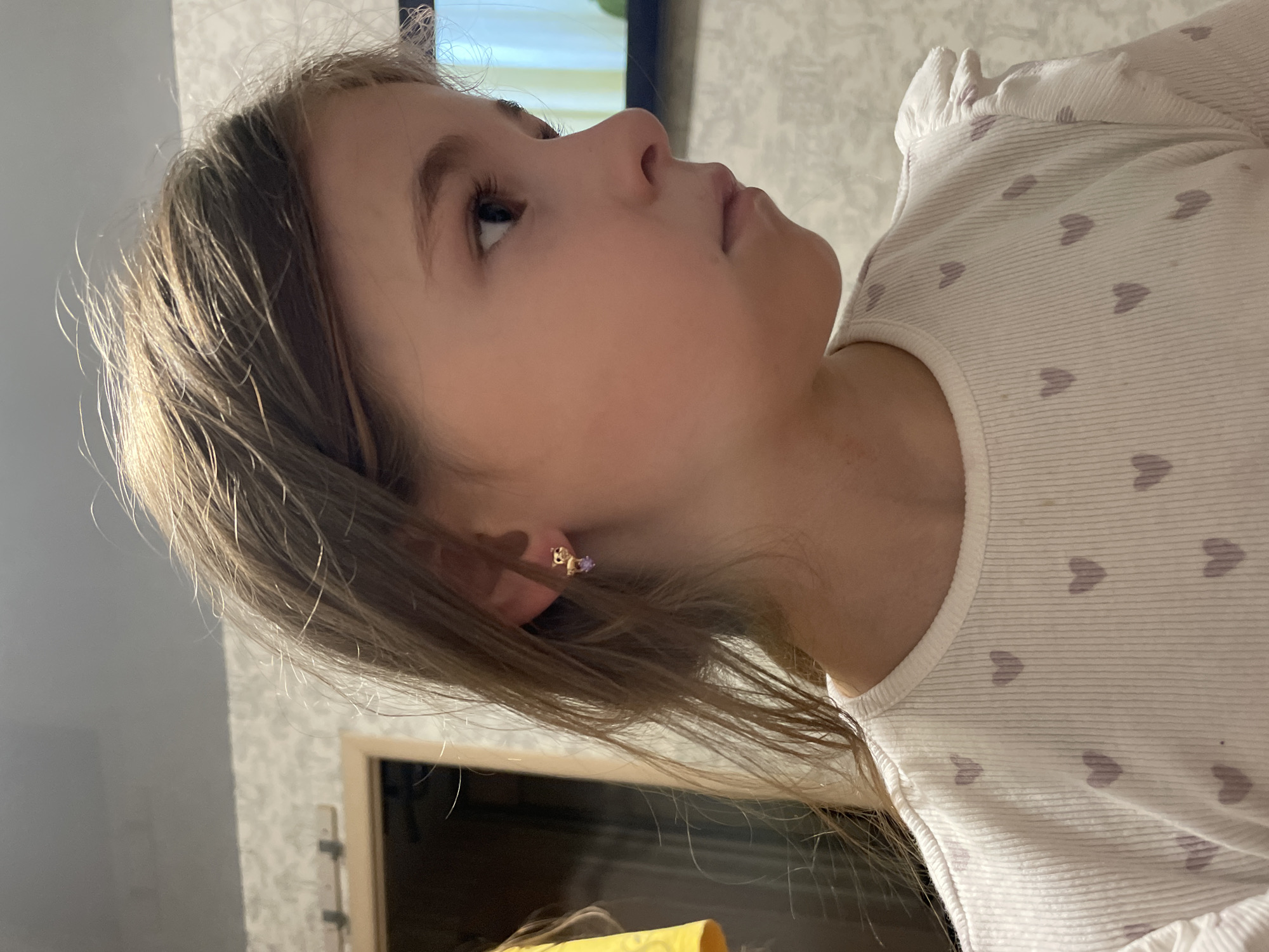 A profile view of a young girl with light brown hair is captured indoors. She is looking to her right. She wears a white ribbed shirt adorned with small purple heart patterns. A delicate gold earring with a purple charm is visible in her left ear. The background consists of a wall with a subtle patterned wallpaper and a doorway. The lighting is artificial and appears to be from an indoor source, casting soft shadows. There is no indication of the city of Sarapul, Russia, in the image or its context.