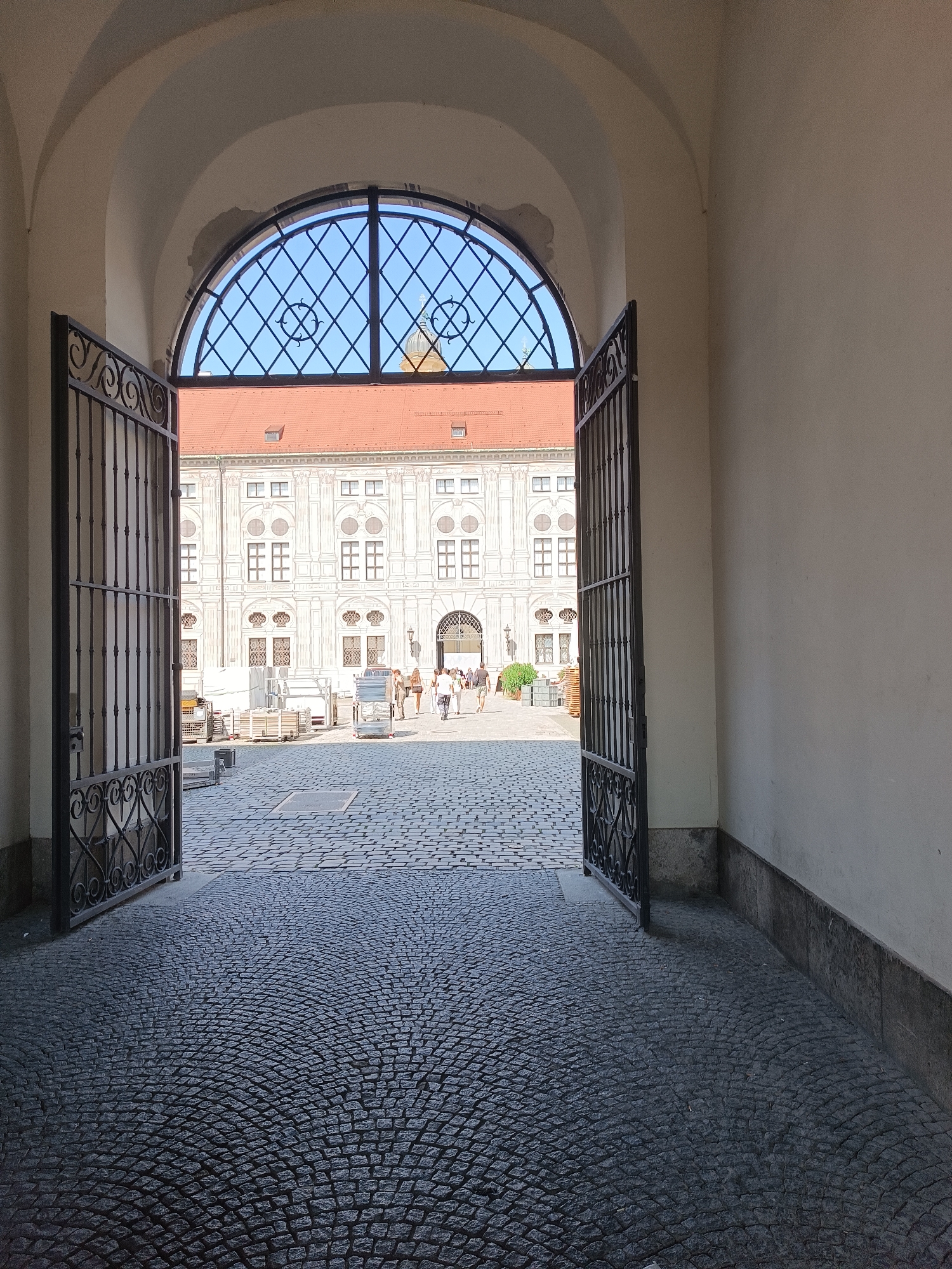 The image depicts a view through an open ornate gate into a sunlit courtyard in Munich, Germany. The courtyard is paved with cobblestones arranged in a fan pattern. In the background, a large, white building with a red tile roof features symmetrical windows and architectural details. A rounded window with a diamond-patterned grate is visible above the building's facade. Several people are seen walking in the courtyard, and some construction materials are stacked near the building. The perspective is from within a shaded archway, with the open gate framing the view of the courtyard and the building beyond.