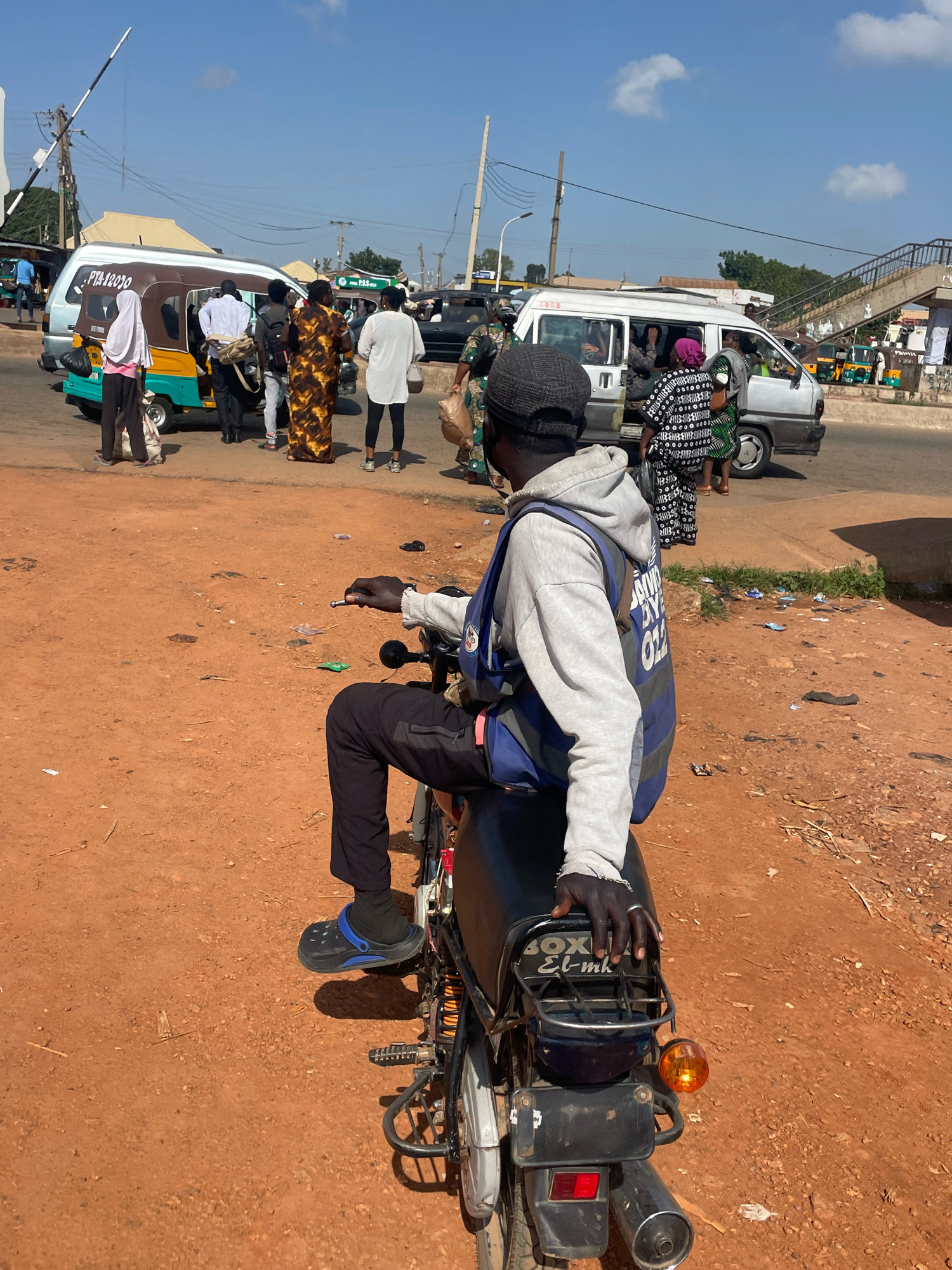 A man is sitting on a motorcycle facing away from the camera, with a bright blue vest over a light gray hoodie. He is wearing black pants and blue and black clogs. The motorcycle is black and silver. The man's right hand is on the motorcycle's handlebars, and his left hand is resting on the seat. The background shows a busy street scene in Gura Top, Nigeria. There are several vehicles, including a white minibus and a yellow and turquoise trishaw. People are standing around, some appearing to be waiting for transportation. The ground is dirt and appears dusty. The sky is blue with a few white clouds.