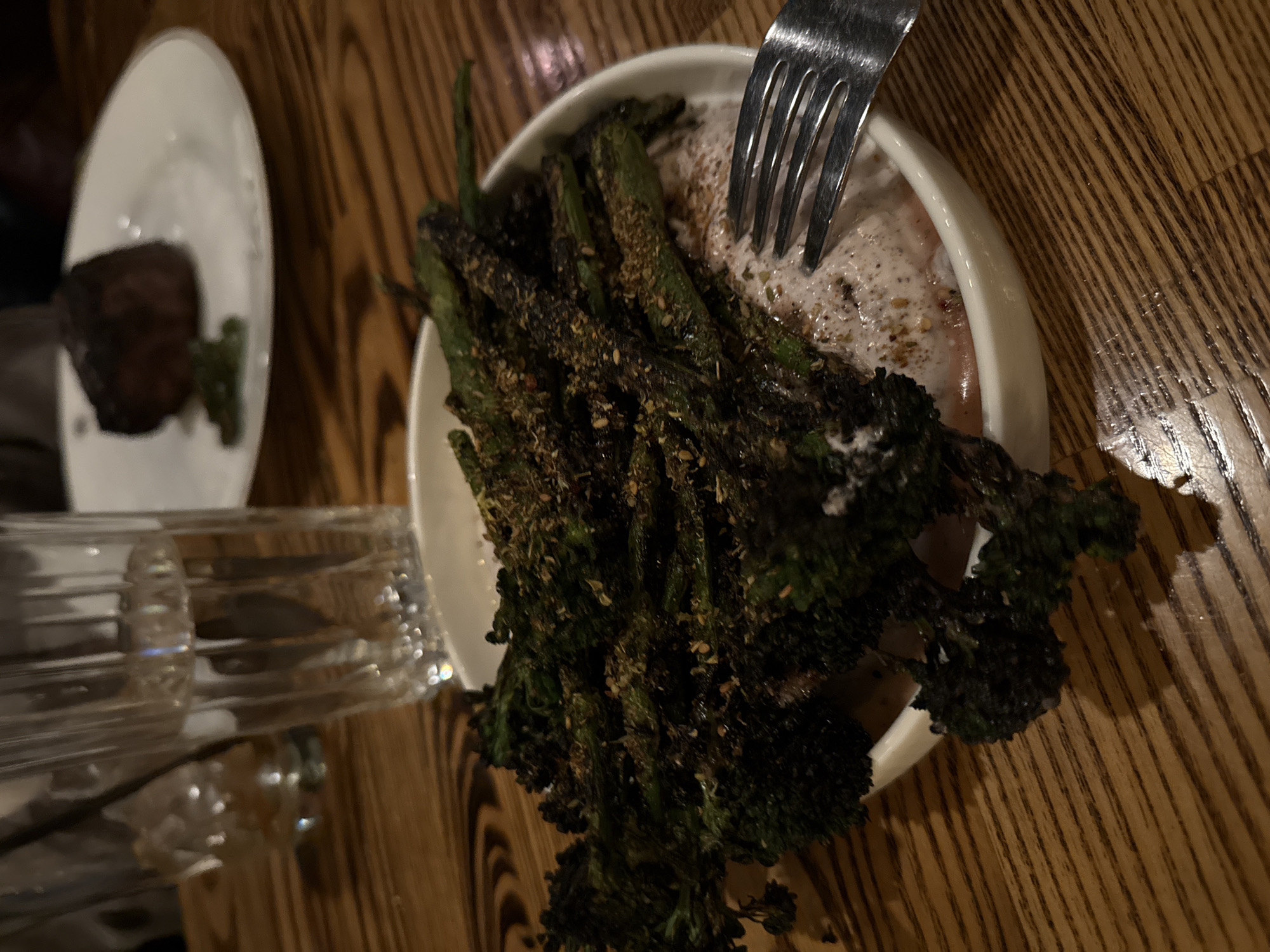 The image depicts a meal at an unidentified location in San Diego, United States of America. A plate containing grilled broccolini seasoned with spices sits in the center of the frame, accompanied by a creamy sauce, possibly hummus or tahini-based. A fork is partially visible, indicating active consumption of the dish. In the background, a second plate holds a small piece of meat alongside a green garnish. The overall lighting is dim, creating shadows and emphasizing the texture of the food. The setting appears to be a restaurant, given the wooden table and tableware. A glass with liquid is also visible.
