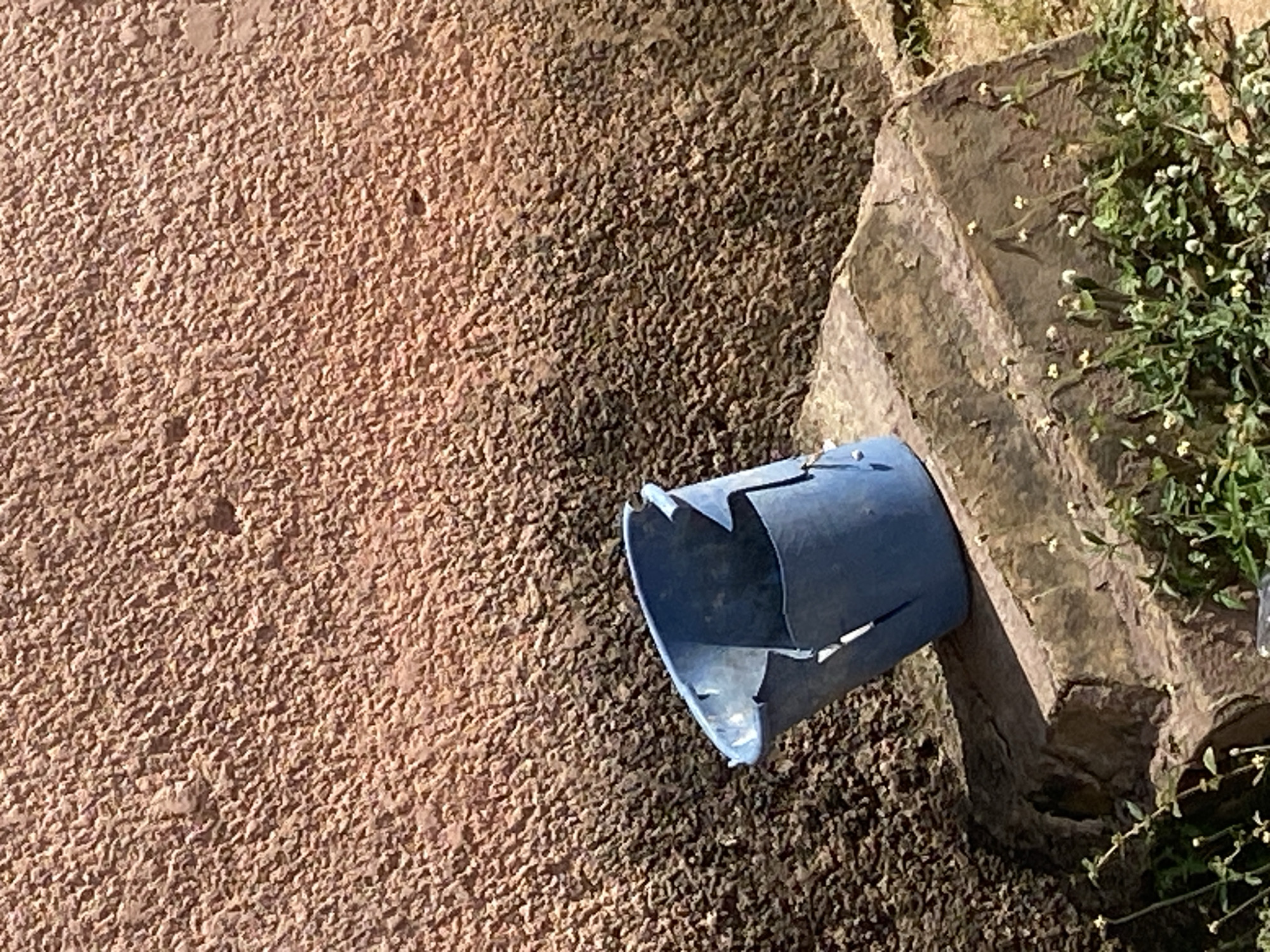 The image shows a close-up of a damaged blue pipe resting on a rough, textured surface, possibly gravel or dirt. The pipe appears to be made of plastic and has several cracks and torn edges, indicating it is broken or deteriorated. To the right of the pipe, there is a large, rough-hewn stone block with some greenery growing around and on top of it. The plants have small white flowers. The background consists of a textured, reddish-brown wall, possibly stucco or concrete, with a very rough, granular finish. The lighting suggests it is daytime, with shadows indicating sunlight. There is no visible text in the image. The scene appears to be outdoors, possibly in a garden or a construction area, given the presence of the pipe, stone, and gravel. There are no people or animals visible. The overall impression is one of disrepair or abandonment.