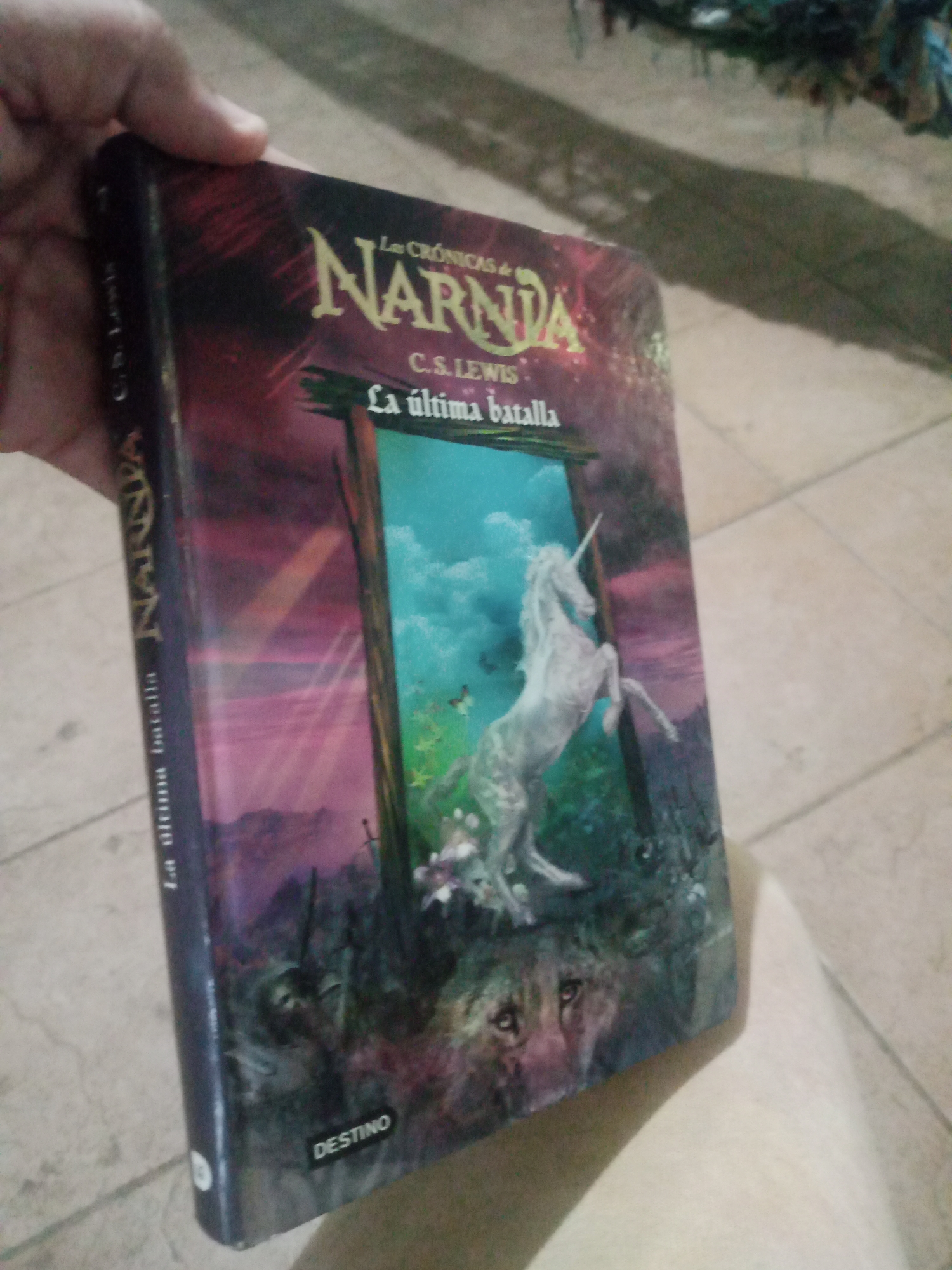 A close-up shot shows a person's hand holding a book titled "Las Crónicas de Narnia: La última batalla". The book cover features a unicorn standing in a mystical landscape with a stormy sky and a wolf-like creature's face in the foreground. The book is oriented vertically and is held at an angle. The background is a tiled floor. The image appears to be taken indoors or in a covered outdoor space. There is no discernible information about the city of Santa Ana, Venezuela, in the image.