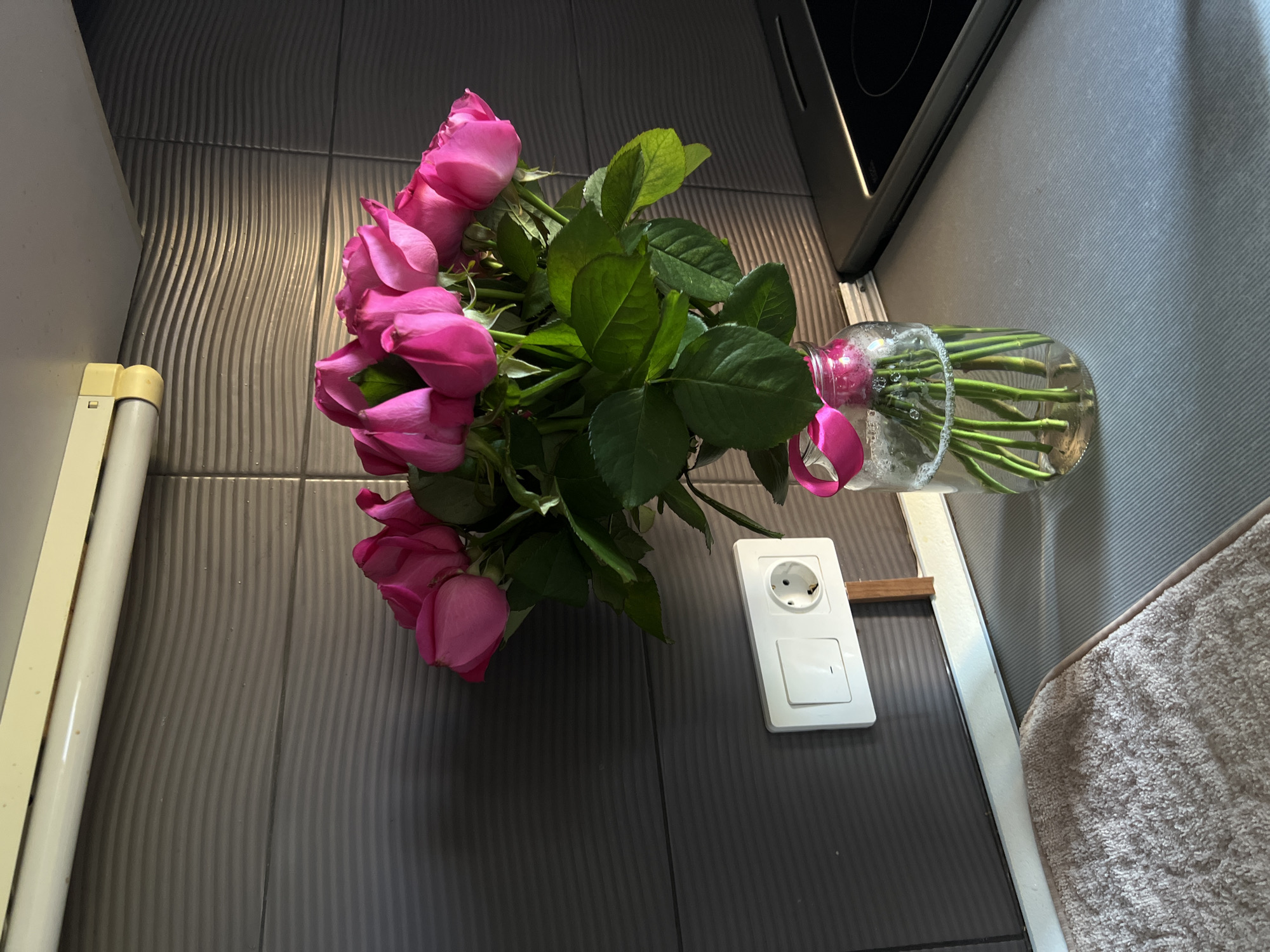 A bouquet of vibrant pink roses sits in a glass jar on a kitchen counter. The roses are tightly packed, their petals unfurled, with lush green leaves fanning out around them. A bright pink ribbon is tied around the neck of the jar, adding a decorative touch. The jar, filled with water, displays the rose stems reaching down to the bottom. Behind the flowers, a dark gray tiled wall serves as a backdrop, with a white electrical outlet and switch mounted on it. Above the tiles, a cream-colored ledge with a fluorescent light fixture is visible. To the right, a portion of a black stovetop is present. In the foreground, a textured beige mat lies on the counter. The scene is brightly lit, with sunlight casting shadows that highlight the textures of the objects.
