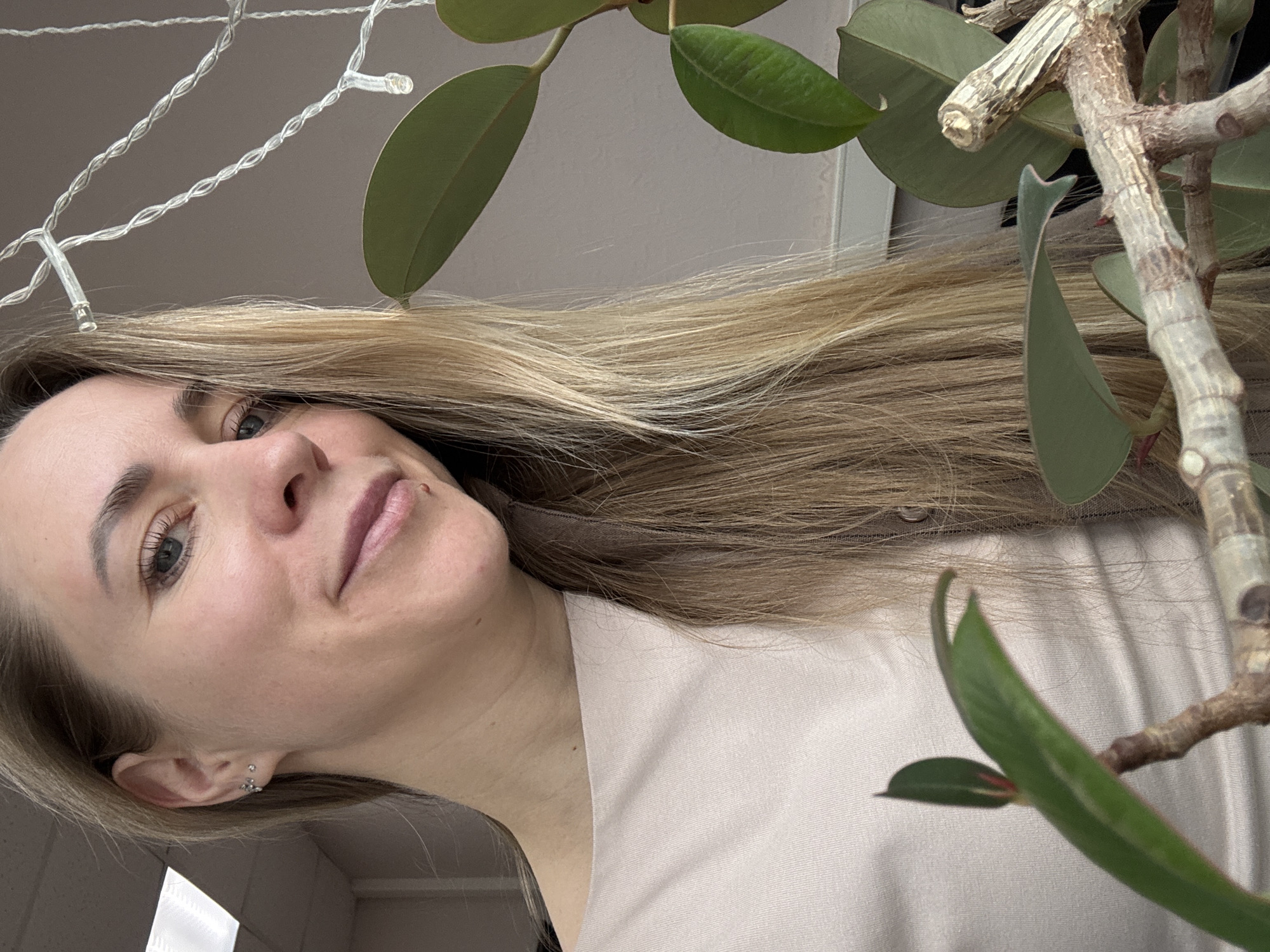 A woman with blonde hair, fair skin, and blue eyes is pictured in a close-up shot. She appears to be smiling slightly. Her attire includes a light-colored shirt. Behind her, a plant with green leaves is visible. In the background, there are some string lights. There is no information that can be directly used to geolocate the location in the city of Chaikovsky, Russia.