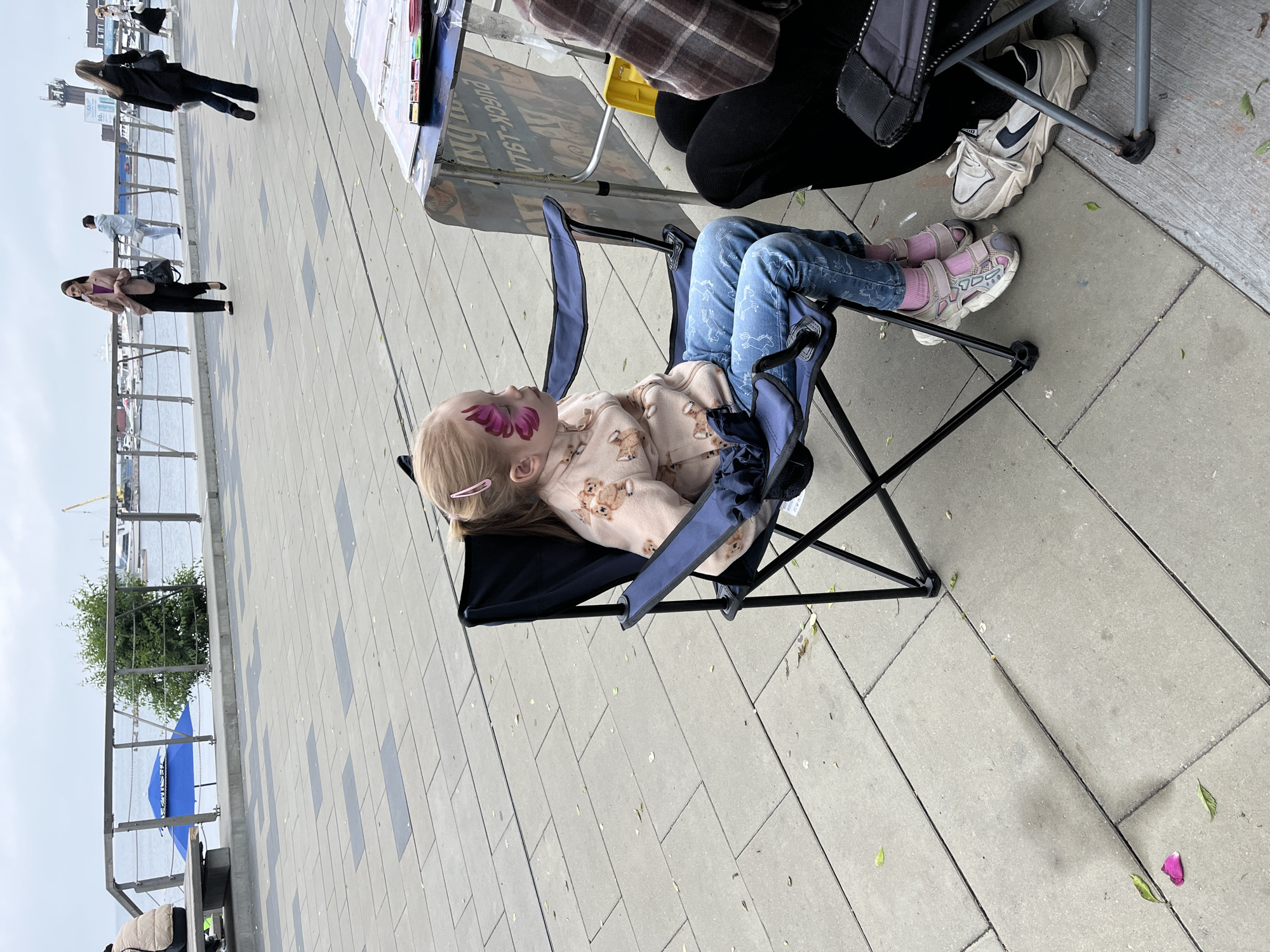 A young girl with face paint, depicting a pink butterfly on her left cheek, sits in a blue folding chair. She wears a light-colored hoodie with a fox pattern and blue jeans. Her feet are clad in pink socks and grey athletic shoes. Beside her, another person, partially visible, sits in a chair. In the background, a paved walkway leads to a railing overlooking a body of water. Several people are visible walking along the walkway in the distance, and boats are moored in the harbor. The scene appears to be set in Vladivostok, Russia, possibly along the waterfront promenade near the Zolotoy Rog Bay. The sky is overcast, and the ground is paved with grey tiles and scattered with fallen leaves.