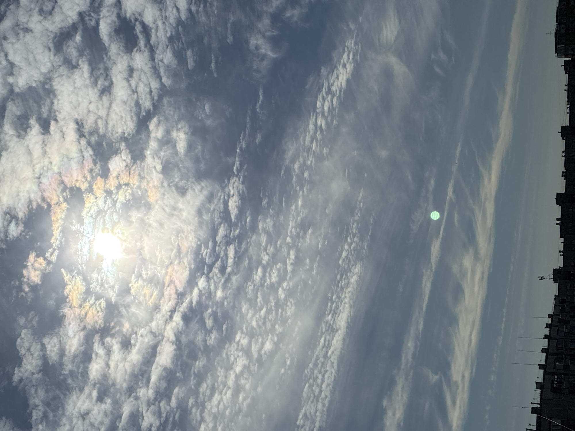The image captures a sky filled with scattered clouds, some of which exhibit iridescence, suggesting a sun pillar phenomenon. The sun itself is bright and partially obscured by clouds, casting a brilliant light. In the lower portion of the image, the rooftops of several buildings are visible, characteristic of an urban environment. The perspective is from below, looking up at the sky and the city skyline.