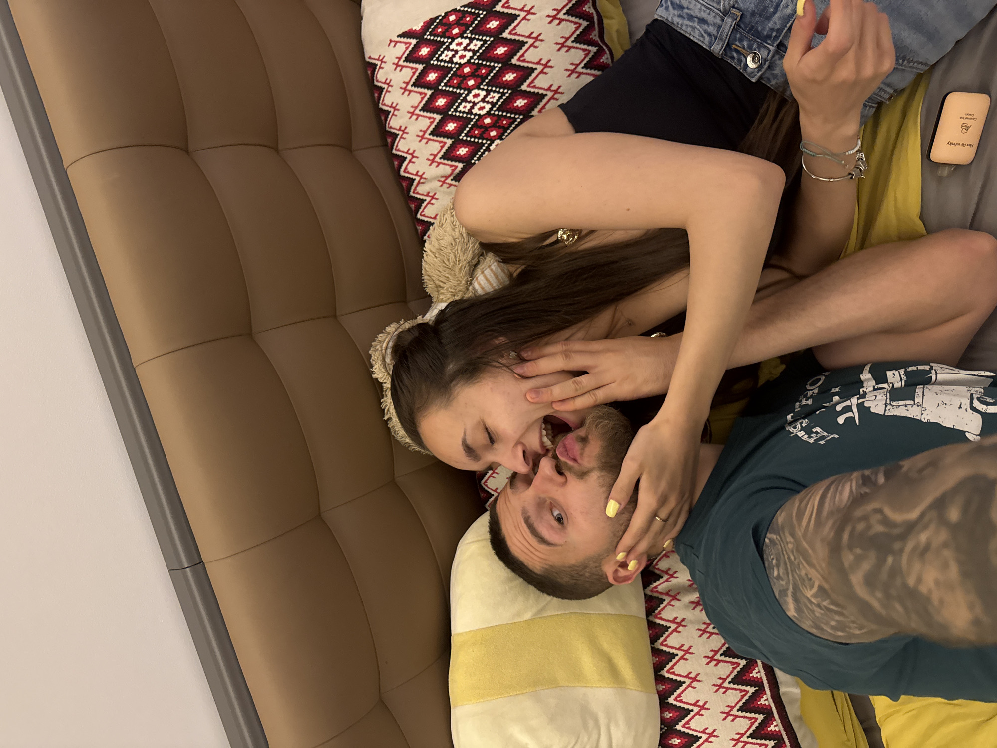 A couple, a woman and a man, are lying on a brown tufted couch with pillows. The woman is wearing a black top and jean jacket, while the man is wearing a green t-shirt. The woman has long dark hair and is making a playful face. The man has short dark hair and a beard. They are touching each other's faces. There is a lotion bottle next to the woman. The pillow has a geometric pattern. The location is most likely in a private home, not specifically identifiable as Sofia, Bulgaria.