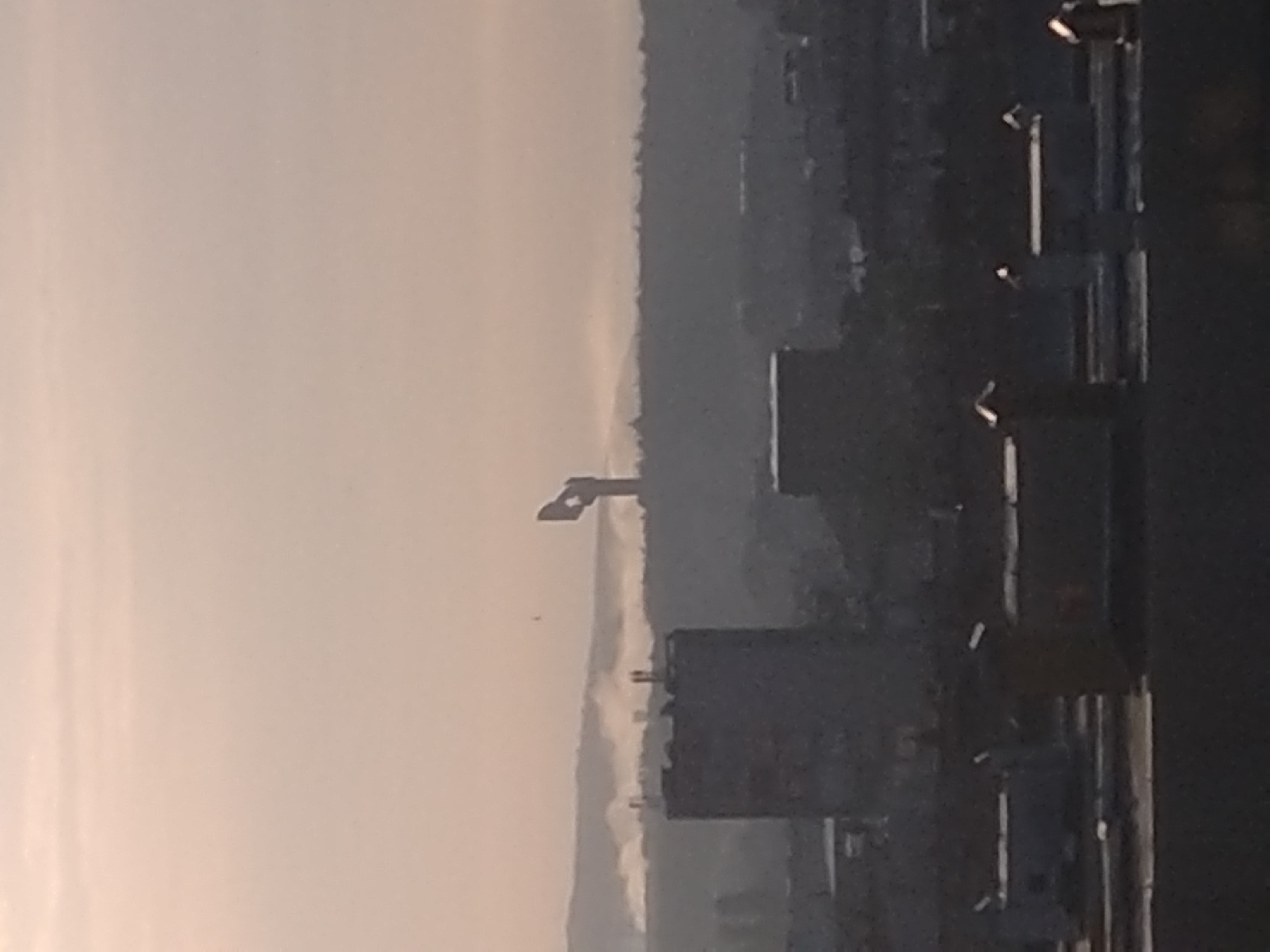 The image captures a distant, hazy view of an urban or industrial landscape, possibly at dawn or dusk. The photo appears to be taken from an elevated position, looking out over a wide area.
In the foreground, on the right side of the frame, are dark, indistinct outlines of buildings and structures, possibly industrial facilities or commercial complexes. These forms are silhouetted against the lighter sky and distant landscape, making details difficult to discern. Several vertical elements, which could be poles, pipes, or parts of building facades, are visible, along with a few bright, reflective spots that might be lights or metallic surfaces catching the ambient light.
Towards the center-left of the image, a prominent, dark, angular structure stands out. This object strongly resembles a crane or a large piece of industrial machinery, characterized by its 'L' or similar bent shape. Below this structure, another cluster of dark, boxy shapes suggests more buildings or industrial components. A tiny, dark speck, possibly a bird or a small distant object, is visible near the crane-like structure.
The background features a range of hills or mountains, appearing soft and muted due to significant atmospheric haze or distance. The upper portion of the sky is a uniform, pale greyish-beige, indicating overcast conditions or a heavy haze layer, contributing to the overall soft and desaturated color palette of the scene.
The lighting suggests either early morning or late afternoon, with soft, diffused light rather than direct sunlight. The pervasive haze creates a subdued and tranquil atmosphere. There is no discernible human activity or visible text within the frame. While the location context is provided as Stara Zagora, Bulgaria, there are no explicit visual cues within the image itself to confirm this.