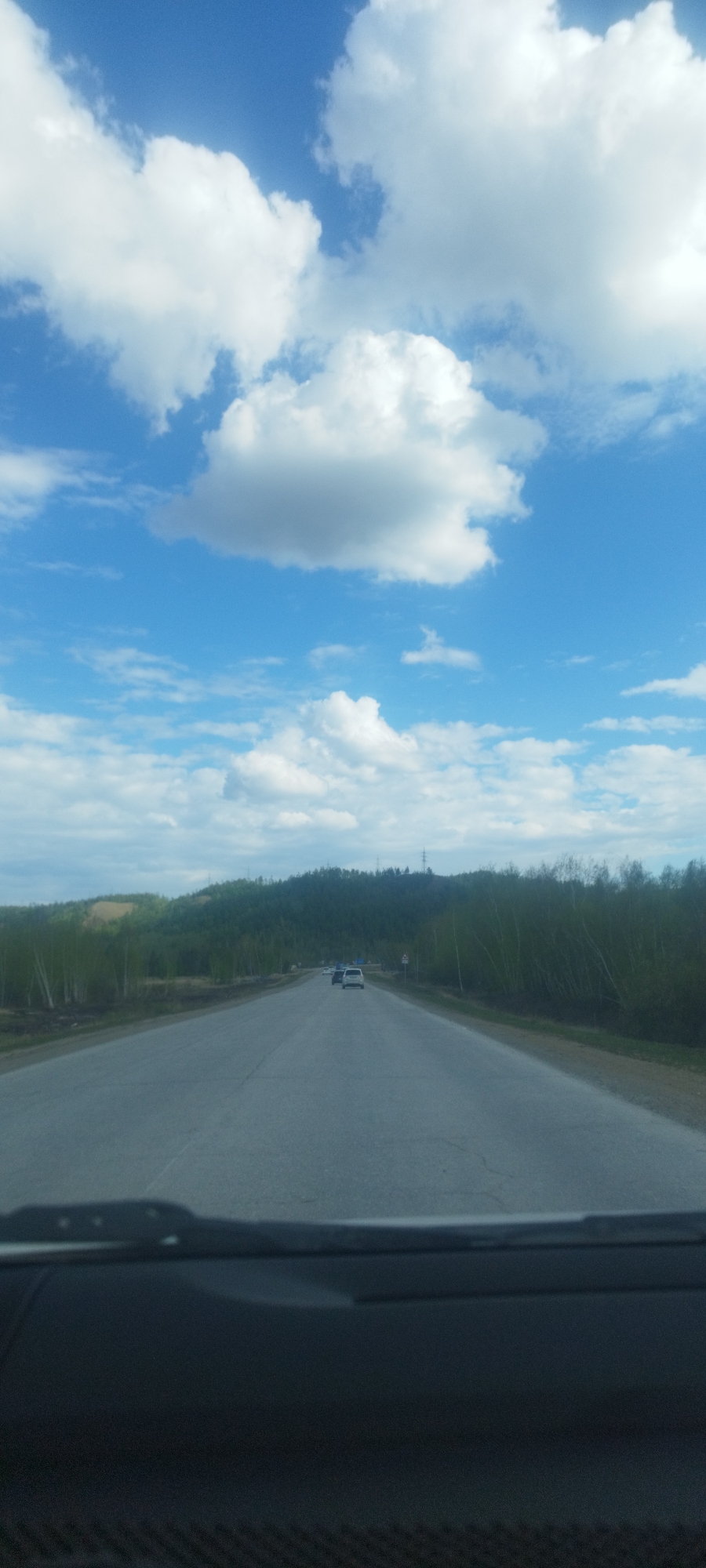 The image shows a road view from inside a vehicle, likely the driver's perspective. The road stretches into the distance, flanked by trees. In the background, there is a hill. The sky is blue with white clouds. Two vehicles are visible on the road ahead. The location is Tabaga, Russia.