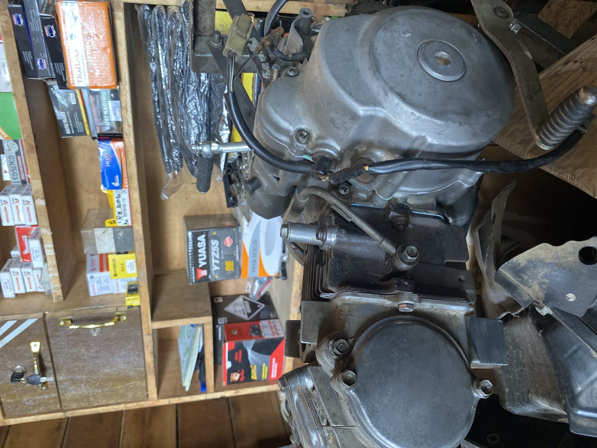 A close-up, eye-level shot captures a motorcycle engine, primarily the crankcase and cylinder head, positioned in the foreground of a cluttered workshop or storage area. The engine appears to be made of aluminum and steel, showing signs of wear and dust accumulation. Various wires and cables are connected to the engine, some neatly tucked, others exposed. A black, rubber-gripped lever is visible to the right of the engine, likely a kickstarter.
In the background, shelves are filled with various items. To the left, a wooden cabinet with two drawers and a key lock is present. The shelves above the engine hold an assortment of boxed motorcycle parts, including what appear to be spark plugs, filters, and engine components. Brands such as "YUASA YTZ5S" and "KOSEIKO" are visible on some boxes. To the left of the engine, on a lower shelf, a red and black box is partially visible. The overall environment suggests a functional, if disorganized, automotive repair or parts storage setting. The image was taken in Bayu, Indonesia.