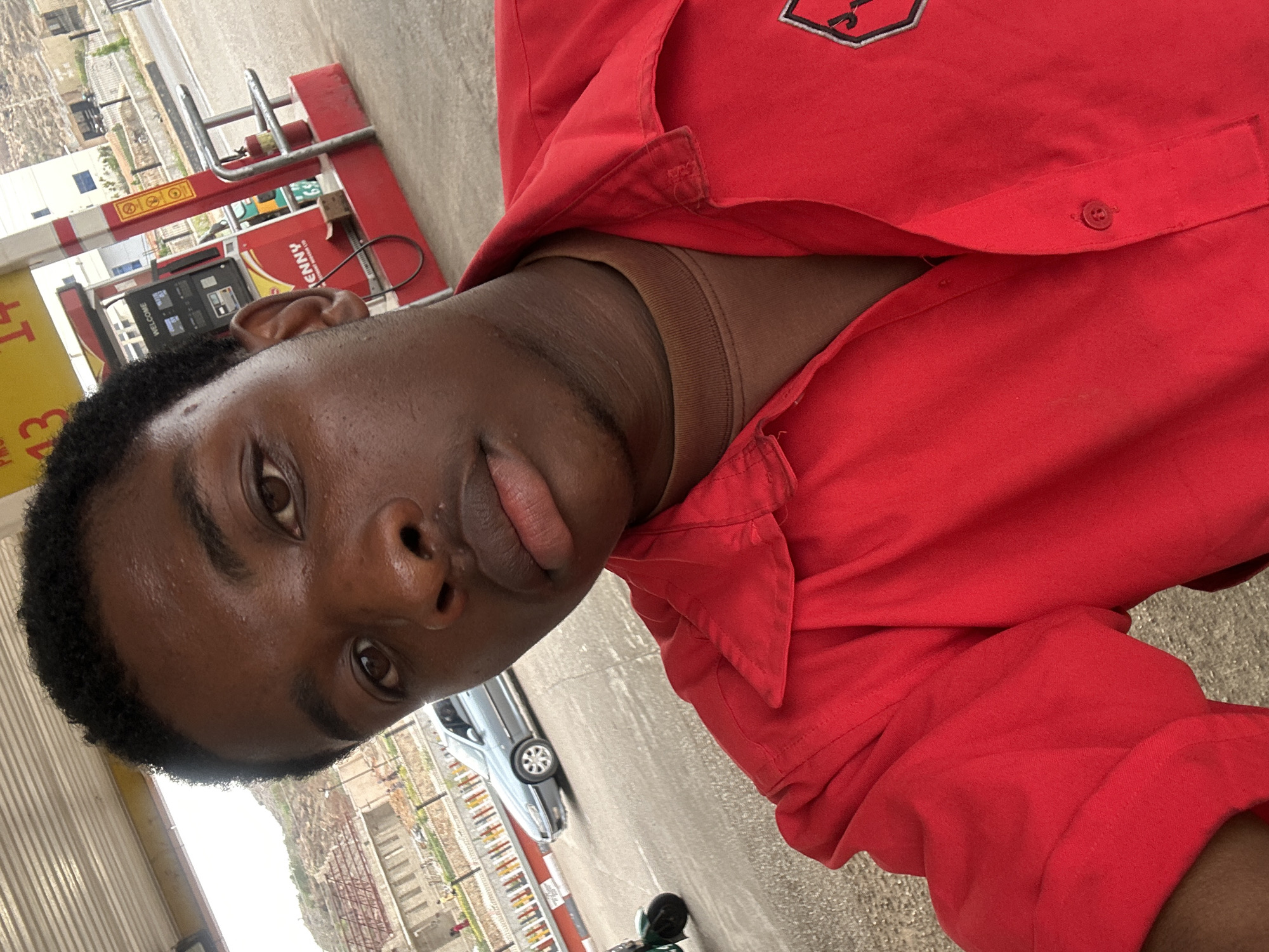 The image is a portrait of a young Black man, captured in an upward-angled selfie, set at a gas station in Jos, Nigeria. The man, with dark skin, short dark curly hair, and dark eyes, looks directly at the camera with a neutral expression, his tongue slightly visible between his lips. He is wearing a red collared shirt, possibly a work uniform, with a dark brown undershirt. A partially visible black and white embroidered emblem, shaped like a shield, is on the left chest of his red shirt.
In the background, a red and yellow fuel pump is clearly visible, displaying the brand name "KENNY" and the word "WELCOME" on its digital screen, along with partial numbers like "659", likely indicating fuel prices. A red and white barrier structure stands nearby, and a coiled black hose or cable is on the ground. A silver car is parked in the mid-ground, and a smaller black object, possibly a scooter, is on the paved ground. The outdoor setting is brightly lit, indicating daytime. Partial text, including "4T" and "43", is visible on a yellow sign in the background.
