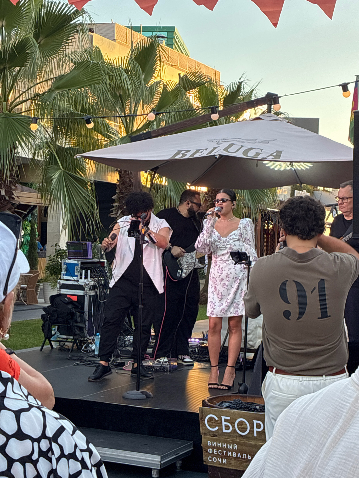The image depicts an outdoor performance stage in Sochi, Russia, with palm trees and a building in the background. A band is performing, with a female singer in a floral dress at the microphone, accompanied by two male guitarists and a drummer. Several people are observing the performance from the foreground, including a person wearing a t-shirt with the number "91" on the back. A wooden barrel with the text "СБОР ВИNNЫЙ ФЕСТИВАЛЬ СОЧИ" (Collection Wine Festival Sochi) is visible on the stage. The overall scene suggests a lively outdoor event, possibly a music festival.