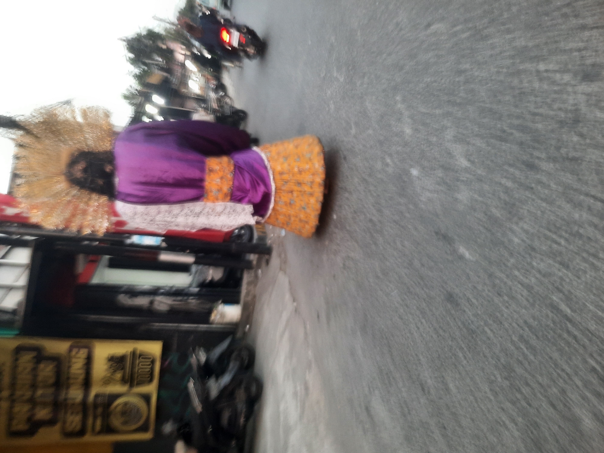The image shows the rear view of a person in traditional attire, possibly a dancer or performer, with a large, elaborate headdress. They are wearing a purple top with an orange sash and a patterned orange skirt. The person is standing on a paved street in what appears to be a commercial area. In the background, there are buildings, motorcycles parked, and other people, including one riding a motorcycle. The overall scene is somewhat blurred due to motion. Based on visual cues like the architecture and the presence of a banner with Indonesian writing ("Pusaka Kuno Seruling" is visible), the location is identified as North Jakarta, Indonesia, likely on a street with shops or businesses.