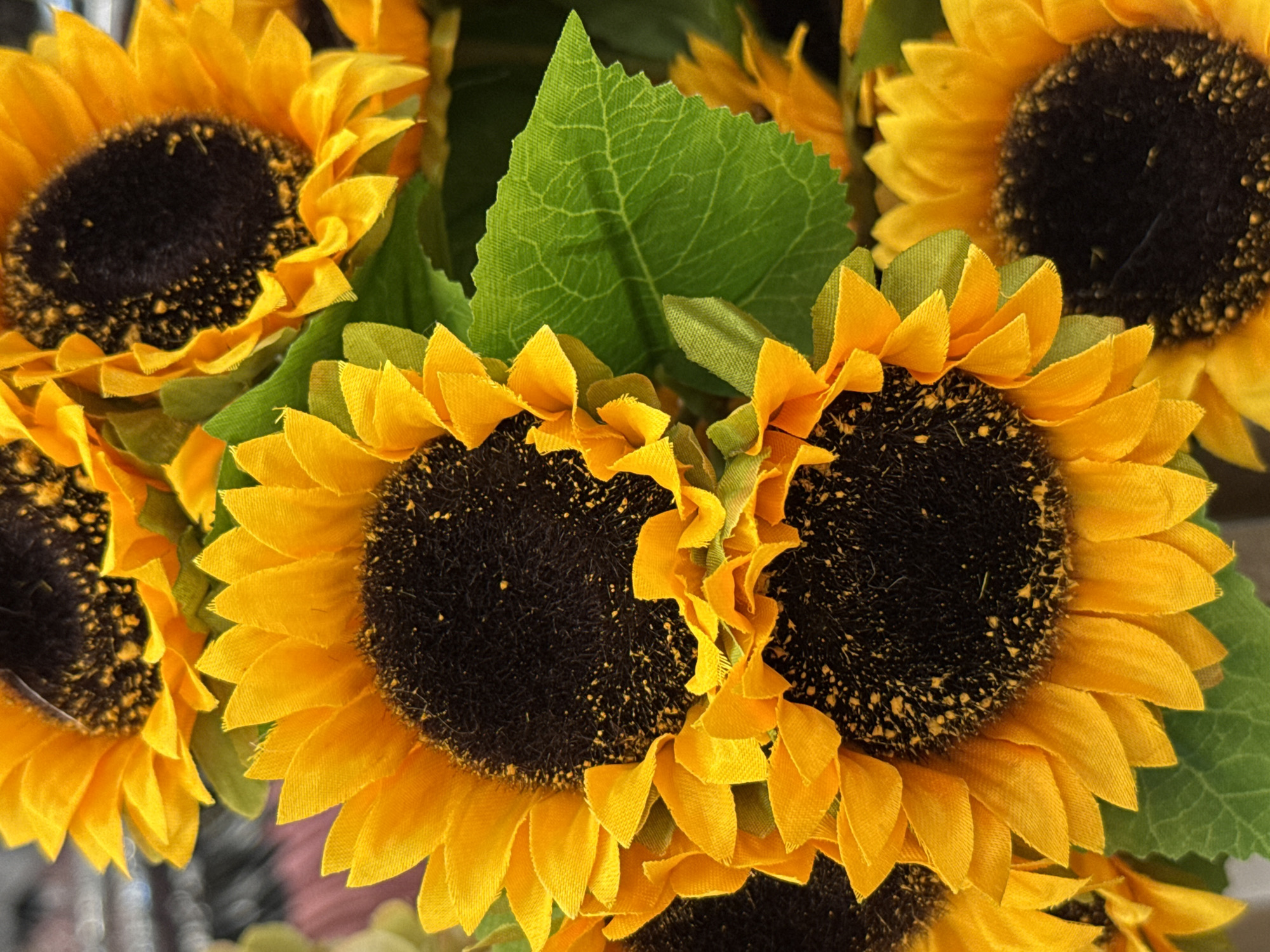 A close-up, overhead shot features a cluster of artificial sunflowers. The sunflowers are a vibrant yellow with dark brown centers speckled with yellow pollen. Green leaves are interspersed among the flowers. The background is blurred and appears to be a retail environment, suggesting these might be displayed for sale. There are no people or animals present. The image is taken indoors, likely in a shop or market. The specific location in Bad Lauterberg im Harz, Germany is not discernible from the image itself.