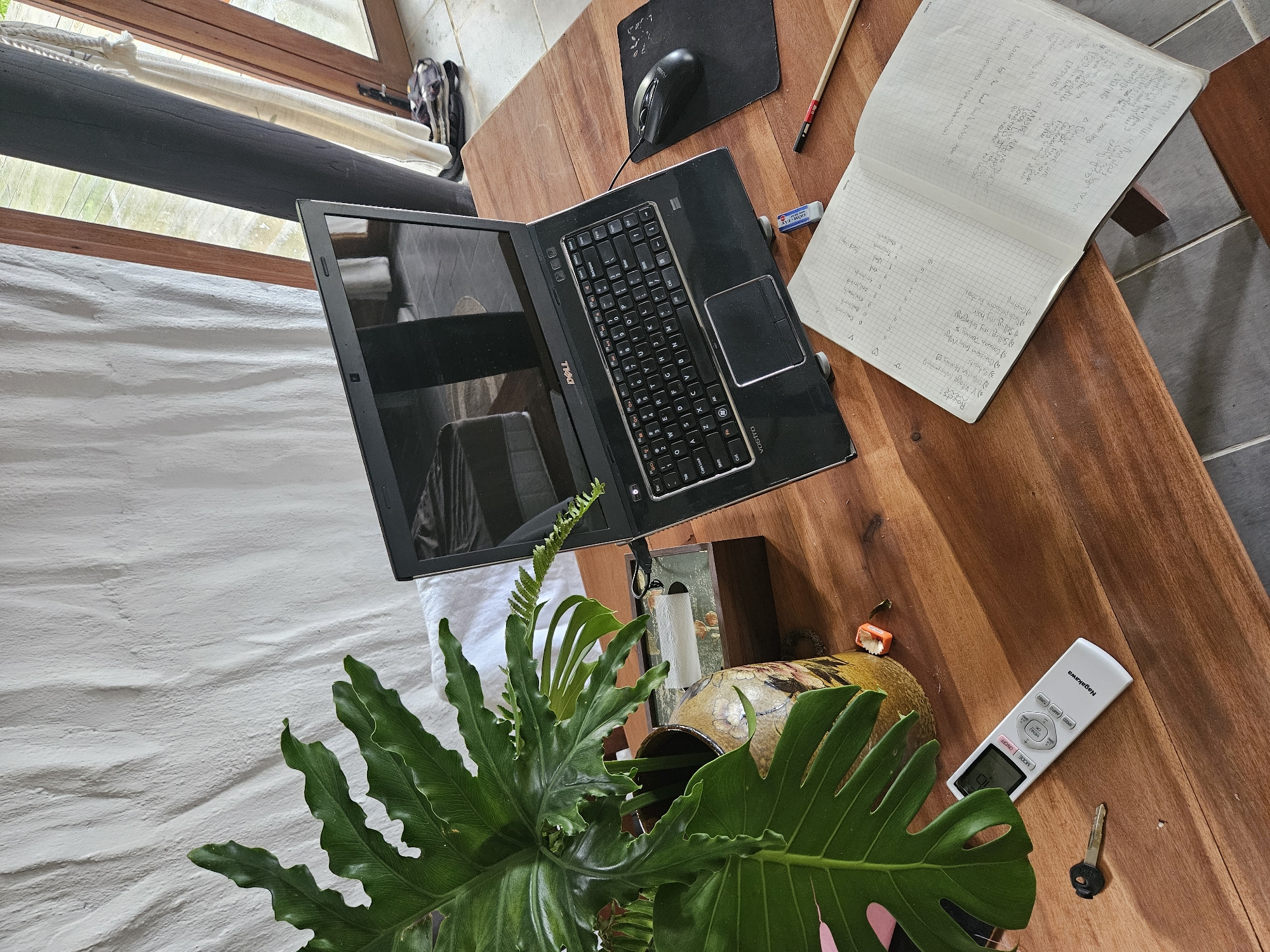 A high-angle, eye-level shot captures a wooden desk set up for remote work, illuminated by natural light filtering through a nearby open doorway and window. The scene is set indoors, likely in a residential or co-working space in Đà Nẵng, Vietnam, evidenced by the tropical plant and the open architectural style.
Dominating the frame is a black Dell laptop, open and positioned slightly off-center. To its right, a black mouse rests on a black mousepad. A partially filled, open notebook with grid paper lies in front of the laptop, its pages covered in handwritten notes in black ink. A single pencil with a red eraser and a small blue eraser are also present on the table.
To the left of the laptop sits a tall, green, leafy plant in a decorative ceramic vase, its variegated pattern in shades of brown and cream. Behind the vase, a small, dark wooden box with a glass lid is visible.
In the foreground, a white remote control with the "Nagakawa" brand name is placed on the desk, alongside a single brass key. An orange object, possibly a small block or toy, is situated near the plant's base.
The background reveals a textured, off-white fabric wall and a glimpse of an open doorway with wooden frames, suggesting an outdoor or transitional space. A dark, cylindrical pillar is also visible, separating two sections of the doorway. The floor appears to be tiled. The overall impression is one of a functional, yet relaxed, workspace.