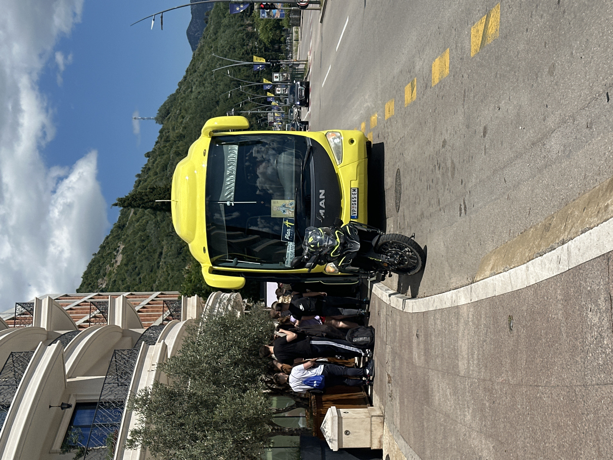 A yellow MAN bus is parked on the right side of the road, facing right. In front of the bus and slightly to the left is a black motorcycle. A group of people, some carrying bags, are standing on the sidewalk to the left of the bus. To the left of the people is a building with balconies and an olive tree. The scene appears to be in Budva, Montenegro, with a lush green hillside in the background and a clear blue sky with some clouds. The road has yellow dashed lines and is moderately busy with other vehicles. The overall lighting suggests it is daytime.