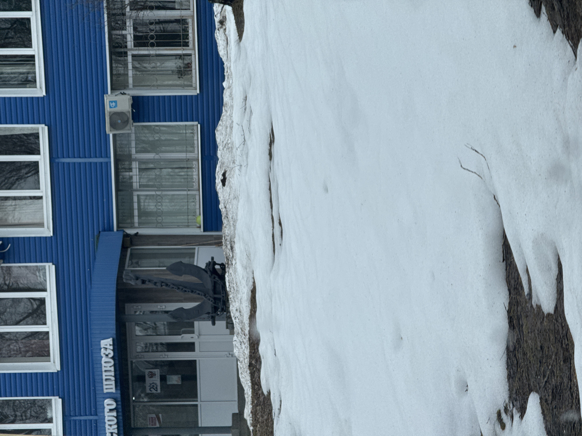 The image depicts a building with a blue facade, partially obscured by a snow-covered area in the foreground. The building appears to have multiple windows and an air conditioning unit. An anchor is fixed above the entrance. The facade indicates that the building is located in Chaikovsky, Russia. The photo appears to have been taken in winter.