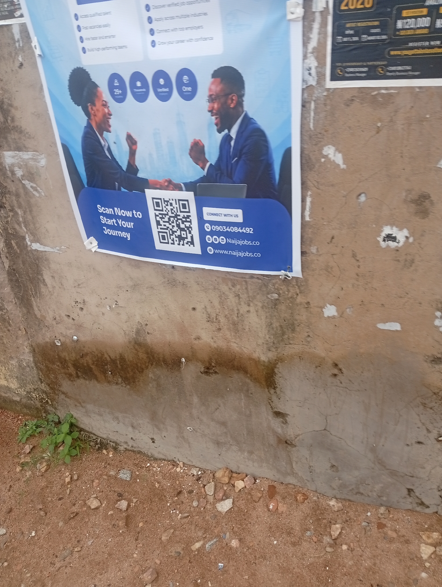 This media file captures a portion of a poster advertising job opportunities, possibly displayed outdoors on a rough, textured wall. Two individuals, a woman and a man, are depicted shaking hands, both smiling and appearing professionally dressed in suits. The woman has her hair styled in a large, elaborate bun, while the man wears glasses. The poster also features icons representing "25+", "Thousands", "Verified", and "One". Below the images, a call to action encourages viewers to "Scan Now to Start Your Journey" with a prominent QR code and contact information for "Naijajobs.co". This includes a phone number, 09034084492, and website addresses.
The poster itself is vibrant with a blue background and white text, contrasting with the weathered, concrete wall it is attached to. The wall shows signs of age, with cracks and patches of discoloration, suggesting it's part of an outdoor urban environment. The ground in the foreground is composed of reddish-brown soil with scattered pebbles and small rocks, and a few sprigs of green vegetation are visible. The lighting appears to be natural, indicating daylight, though no specific time of day can be determined. The overall scene is one of a promotional advertisement placed in a public, somewhat unkempt, location, likely in Jalingo, Nigeria, given the context. The poster's message is clear: it's a platform for job seekers and employers to connect.