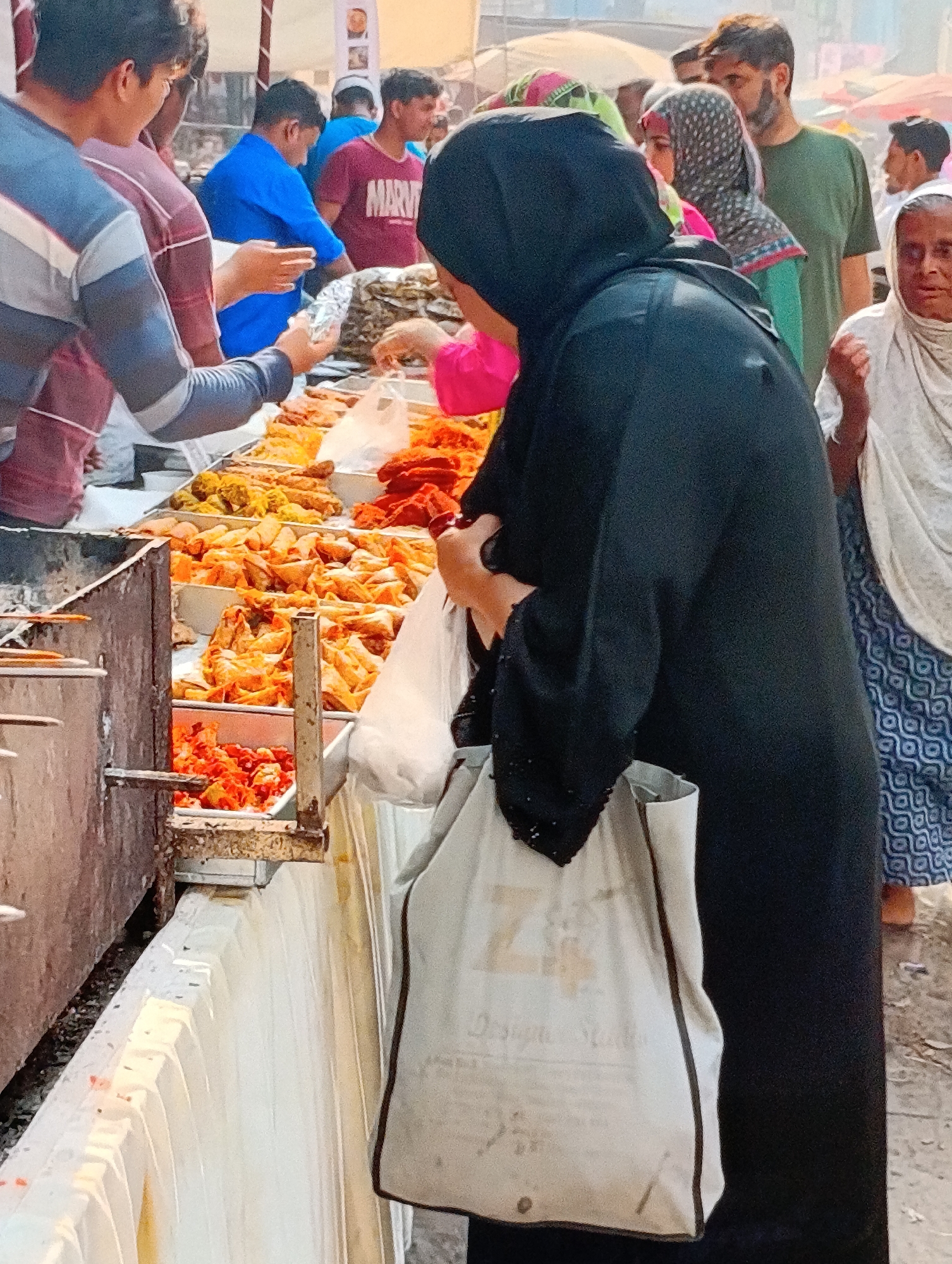 In a bustling outdoor market, a diverse group of people gathers around a street food vendor. The foreground is dominated by a food stall laden with a variety of fried items, including samosas and other unidentified snacks. Several individuals are actively purchasing food. A woman wearing a black hijab and abaya holds a plastic bag in one hand and a shopping bag in the other, suggesting she is a customer. To her right, an older woman in a white headscarf looks on. A man in a green shirt stands behind her, part of the crowd surrounding the stall. The background shows additional market-goers and vendor stalls under the open sky.