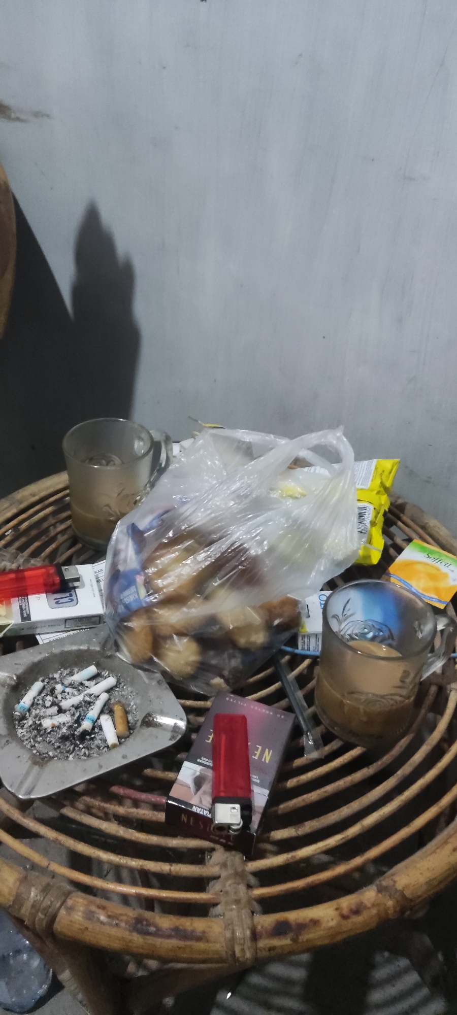 A still life photograph captures a collection of items on a woven wooden surface, likely a table. The scene is lit by ambient light, casting shadows on a textured wall behind. In the foreground, an ashtray is filled with numerous cigarette butts and ashes. Adjacent to it, a pack of cigarettes and a red lighter are visible. Further back, a clear plastic bag contains what appear to be baked goods. Two glasses are present; one is partially filled with a light brown liquid, possibly coffee or tea, and the other is empty or contains a minimal amount of liquid. A small yellow packet, possibly a snack or condiment, is also on the surface. The overall impression is of a casual, lived-in environment, possibly a moment captured at a residence in Karangwangun, Indonesia.