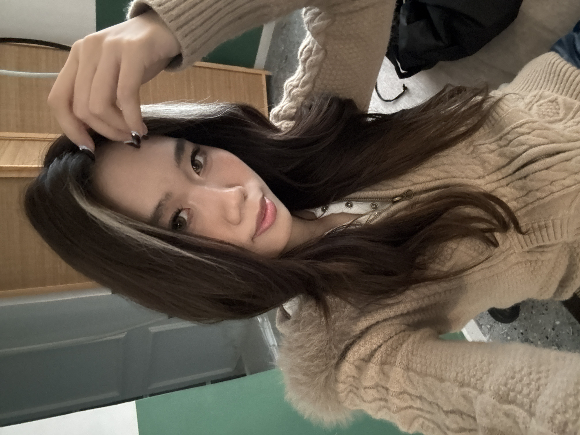 An indoor portrait image depicts a young East Asian female subject in Liangjiang, China. The subject is positioned centrally, facing forward and making direct eye contact with the viewer. She possesses long, dark brown hair with a distinct, lighter-colored streak visible on the right side of her face. Her right hand is raised, with fingertips resting on her forehead; the fingernails are painted dark with metallic silver tips. She is wearing a beige or light tan cable-knit sweater, featuring a prominent faux fur-lined hood. Beneath the sweater, a white collared garment with a partial zipper closure and three metallic snap buttons is visible, along with a metallic chain necklace ending in a circular pendant. The background consists of an interior space featuring a partial green wall section, an adjacent light gray door or panel, and a vertical wooden surface, possibly cabinetry, to the upper right. The floor is visible as a gray speckled surface. A black object, resembling a bag or fabric item, rests on the floor to the subject's lower right.