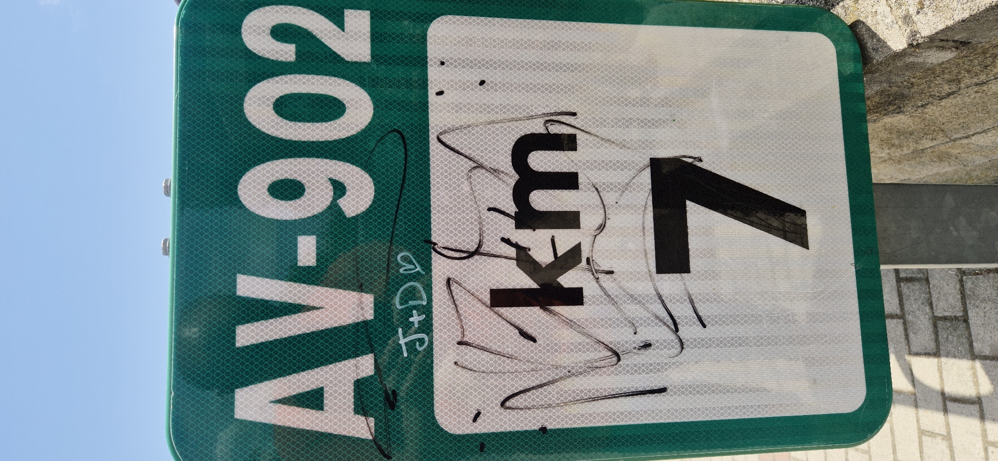 A green and white reflective road sign is mounted on a metal pole. The sign has a green border and displays "AV-902" in large white sans-serif capital letters and numbers. Below this, there is graffiti. To the left of "AV-902", there are the initials "J+D" with a heart symbol written in white marker. The lower portion of the sign is white with a diamond pattern and displays "km" in large black sans-serif capital letters. Below "km", the number "7" is displayed in large black sans-serif numbers. The sign has been vandalized with black spray paint or marker, which covers much of the white lower portion of the sign and partially obscures the "km" and "7". The background shows a bright blue sky and a paved walkway made of brick. To the right of the sign, a stone wall is visible.