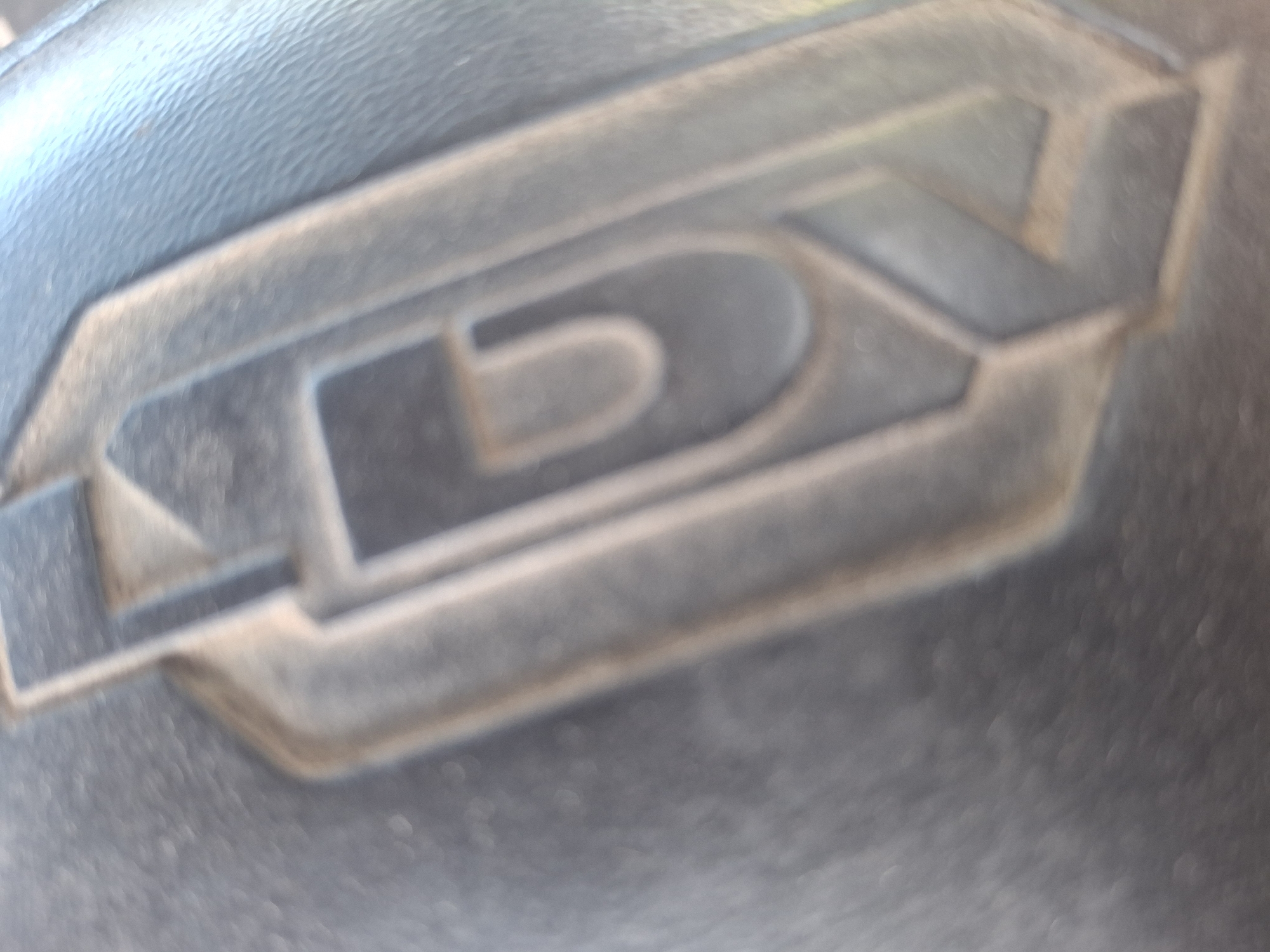 The image shows a close-up, slightly blurred view of a black rubber object with a raised, molded design. The design appears to be a stylized letter or symbol, possibly a logo or branding. The texture of the rubber is visible, with some fine lines and imperfections. The lighting casts shadows within the recessed areas of the design, highlighting its three-dimensional nature. The background is also black rubber, but less detailed due to the focus and blur. The image does not provide any context for the location within the city of Kalush, Ukraine, or any other environmental details. There are no people, actions, or interactions depicted.