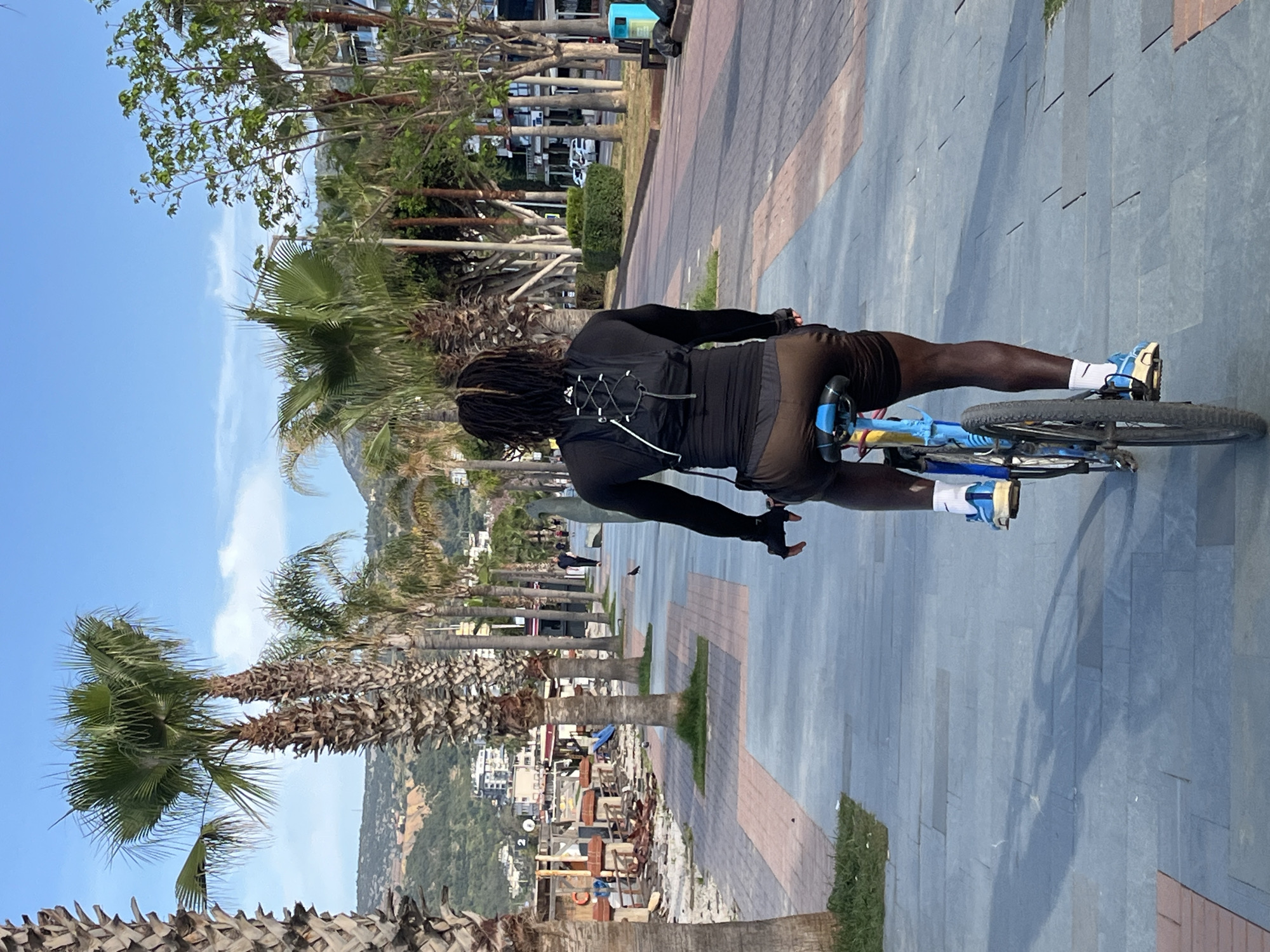 A person is riding a bicycle on a paved walkway in Alanya, Turkey. The person is wearing black long-sleeved clothing, brown shorts, white socks, and blue and white sneakers. The bicycle is blue with black tires. The walkway is made of gray and brown paving stones. Palm trees line the walkway. In the background, there is a mountain and buildings. The sky is blue with some clouds. A few other individuals are walking in the distance.