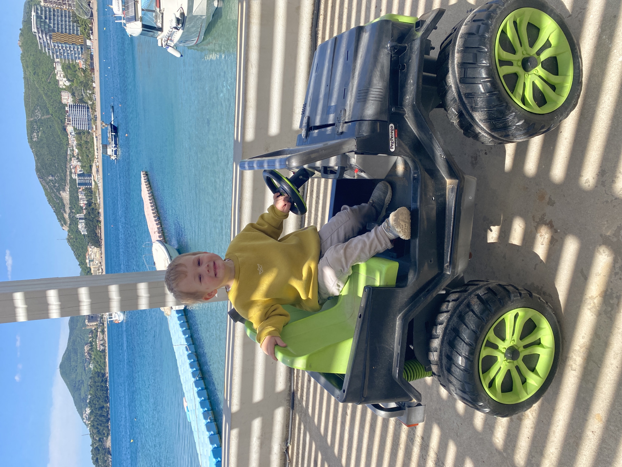 A young child sits in a green and black toy jeep on a sunny day, likely in Rafailovići, Montenegro. The child is wearing a yellow sweater and light-colored pants, and is holding the steering wheel. Behind the child and the jeep is a waterfront scene with blue water, mountains, buildings, boats, and a pier. White railings are also visible in the background. Shadows from a nearby structure cast striped patterns across the ground where the jeep is parked.