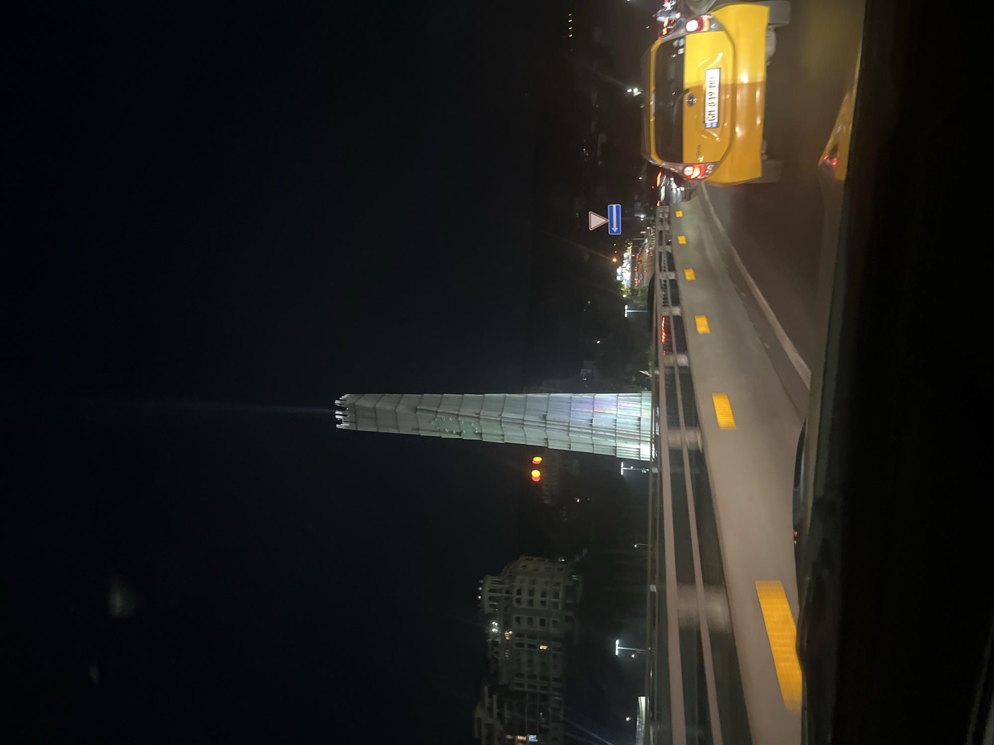 The image captures a nighttime scene in Tbilisi, Georgia, focusing on a modern, illuminated tower that appears to be a landmark. The tower is tall and slender, with a distinct, faceted design, and is lit with a vertical beam of light extending upwards into the dark sky. The tower is surrounded by the city's infrastructure, including a highway with cars and streetlights. On the left side of the frame, a multi-story building with architectural details characteristic of older European buildings is visible on a hill. The foreground is dominated by the view from inside a car, showing the dashboard and windshield, and the rear of a yellow car driving ahead on the highway. The yellow car has a license plate that reads "GM 819 MD". The highway has reflective markers on its barriers, indicating it's a well-maintained road. The overall impression is one of a bustling urban environment at night, with the illuminated tower serving as a central visual element.