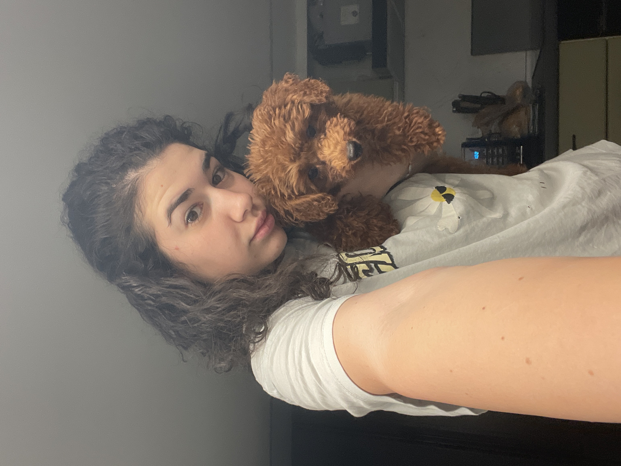 A young woman with curly dark hair is lying on her side, facing the camera. An auburn-colored toy poodle is nestled close to her, seemingly on her chest. She is wearing a white t-shirt with a flower design on it. The background is blurry but appears to be an interior space. The image does not provide enough information to determine the location in Sofia, Bulgaria.