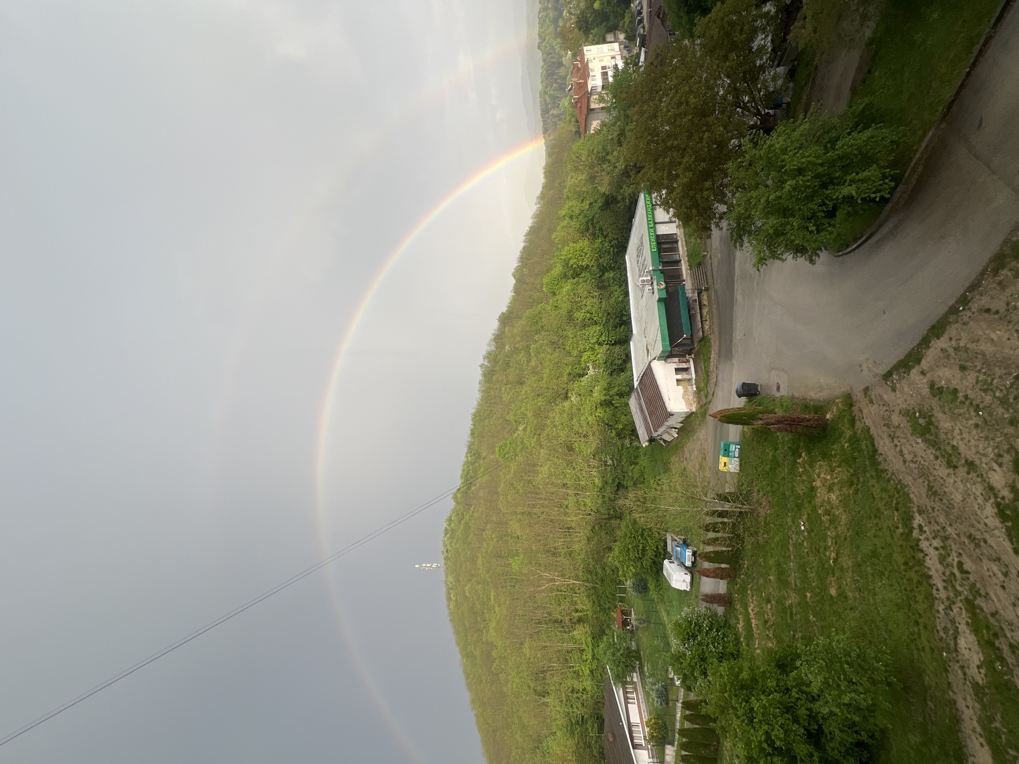 The image shows a partial view of Elena, Bulgaria, under overcast skies and a double rainbow. The foreground features a hillside densely covered with trees, next to buildings and a road. A white and green building is visible near the top of the hill. A white van and a blue van are parked in a grassy area. A power line stretches across the sky near the bottom left of the rainbow.
