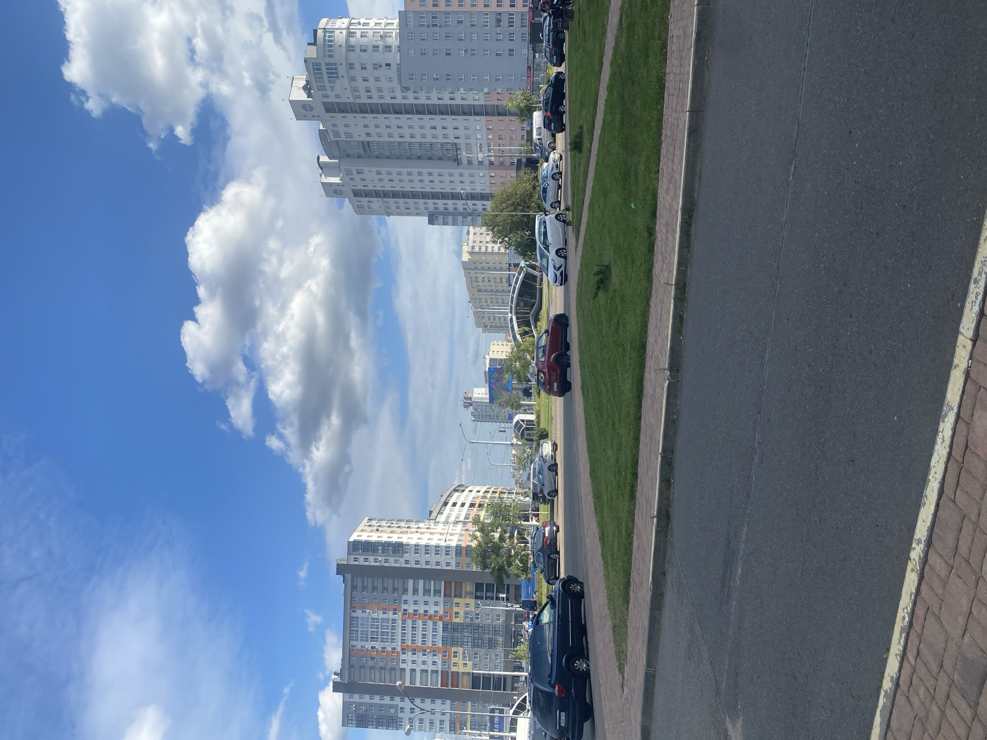 The image captures a wide view of a street scene in Minsk, Belarus, under a bright, partly cloudy sky. In the foreground, a paved road is visible with a distinct red brick sidewalk bordering it on the left. Beyond the road, a circular grassy area with manicured bushes and trees separates it from the traffic lanes.
On the left side of the frame, a tall, multi-story apartment building with a facade featuring a mix of gray, white, orange, and yellow colors stands prominently. Several cars, including a dark blue minivan and a black sedan, are parked or driving in front of this building.
To the right, another large, modern apartment building with a rounded, multi-faceted design and predominantly white and light gray coloring dominates the skyline. Numerous cars are seen on the road in front of this building as well, including a red hatchback, a white sedan, and several other vehicles. In the background, a series of other residential buildings and some streetlights are visible, contributing to the urban landscape. The overall impression is one of a typical residential area in a modern city, with well-maintained buildings and infrastructure.