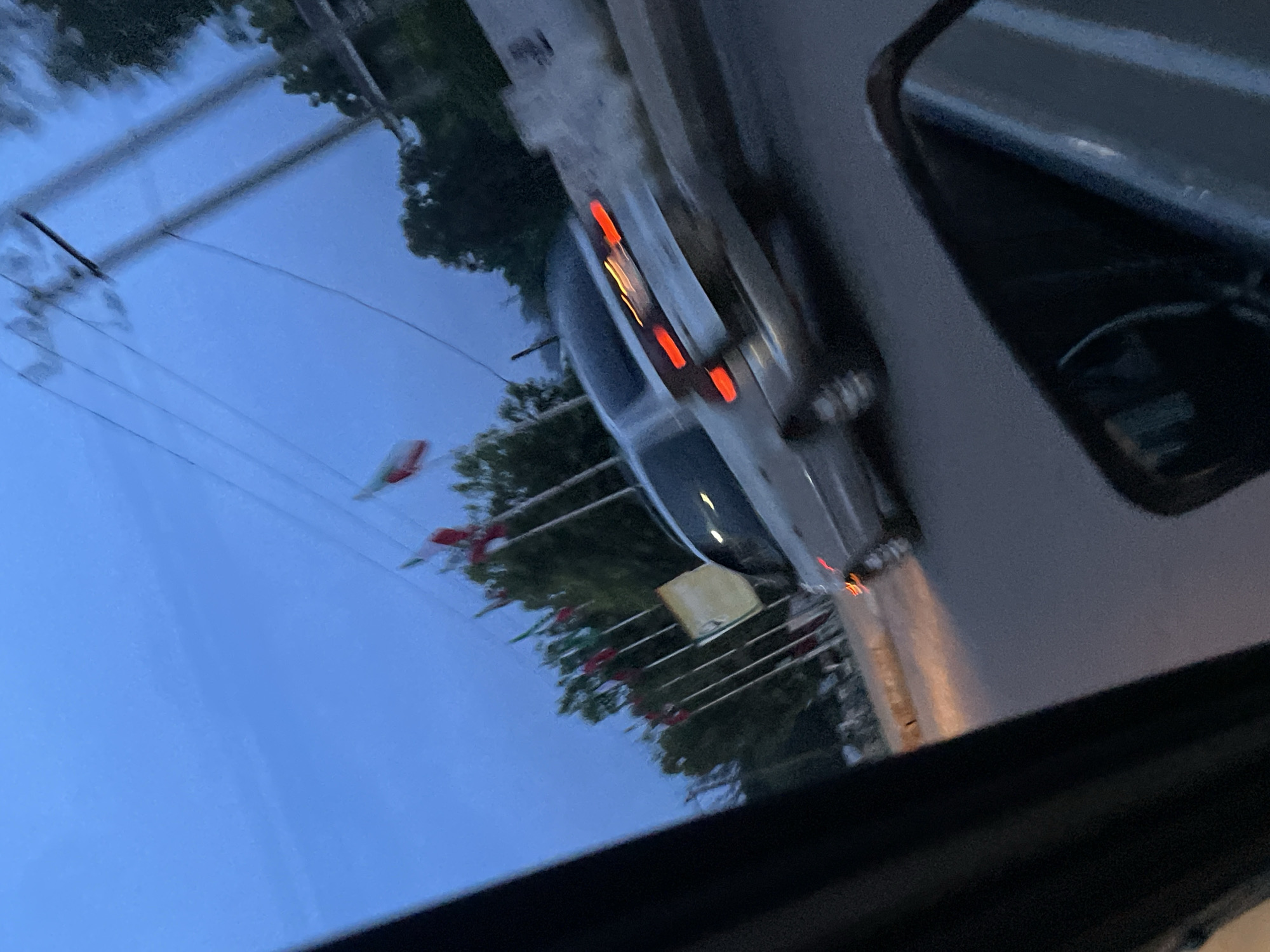 The image shows a reflection of a street scene, possibly taken from inside a vehicle. The dominant subject is a silver sedan with its brake lights illuminated, suggesting it is either stopped or moving. The car is seen from an angle, with its rear end more prominent.
Reflected in what appears to be the car's roof or a window, we see a street with overhead power lines and trees. There are also some flags or banners visible, adorned with red and green elements, adding a splash of color to the scene. A light-colored rectangular object is also present in the reflection, its purpose unclear. The sky appears overcast or it's twilight, indicated by the dim, blueish hue, and the low visibility of natural light.
The overall impression is that of an urban or suburban environment in Jos, Nigeria, during either dawn or dusk. The illuminated brake lights and the general mood of the scene suggest a moment of transition, perhaps a pause in transit. There is no discernible text in the image.
