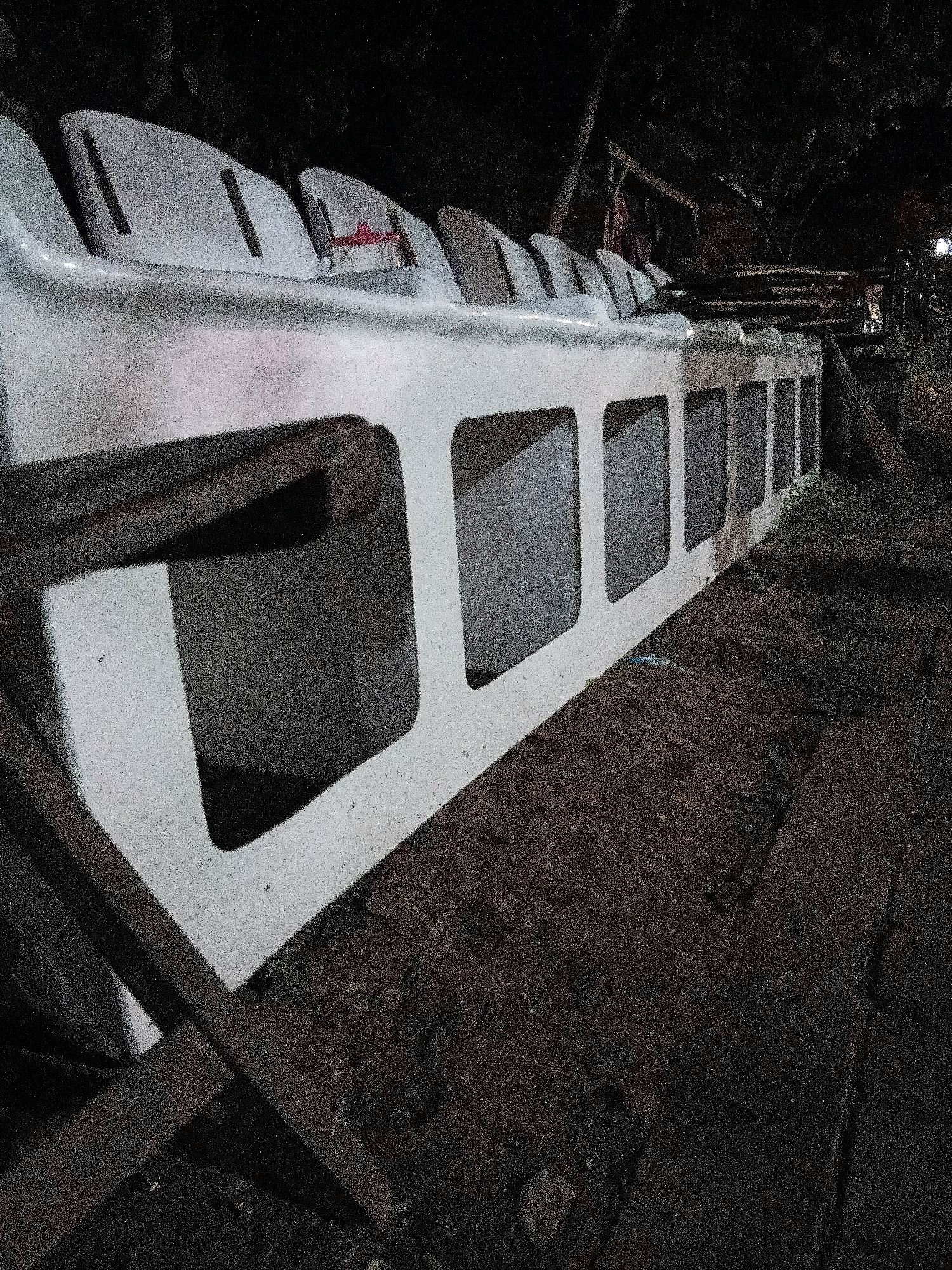 In Cikal, Indonesia, a row of empty, white stadium seats is shown at night, viewed from a low angle. The seats are arranged side-by-side, with a white bench structure supporting them. Each seat has a contoured design and a small, vertical slot on its backrest. The bench has several rectangular openings along its side. The ground beneath the seats appears to be dirt and gravel, with some dark vegetation and wooden supports visible. The background is dark and out of focus, with hints of trees and distant lights suggesting an outdoor location.