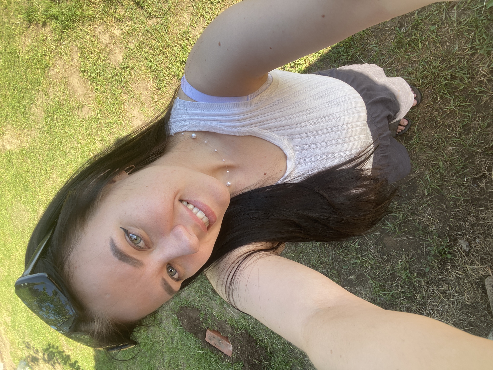 A woman takes a selfie outdoors in what appears to be a grassy area in Chaikovsky, Russia, during the daytime. The image is rotated 90 degrees clockwise, showing the woman's face in the upper left corner and her body extending diagonally across the frame. She has long, dark hair that cascades over her shoulder and arm, visible on the right side of the image.
The woman is smiling and looking directly at the camera. She has light-colored eyes and is wearing eyeliner. A pair of dark sunglasses is resting on top of her head. She is wearing a light-colored, possibly white or off-white, ribbed tank top, and a purple strap, possibly from a bra, is visible beneath her tank top on her left shoulder. A delicate necklace with small pearls is visible around her neck.
Her right arm extends towards the camera, holding the phone. Her torso and legs are partially visible, with a dark skirt or fabric draped around her lower body. Her feet, wearing dark flip-flops or sandals, are visible in the lower right portion of the image, resting on the grass.
The background consists of green grass and patches of dirt, suggesting an outdoor, possibly natural or semi-natural, setting like a yard or park. The lighting indicates it is sunny, as there are distinct shadows visible, particularly on the grass and around her body. A reddish brick is visible on the ground in one of the close-up crops. No visible text is present in the image.