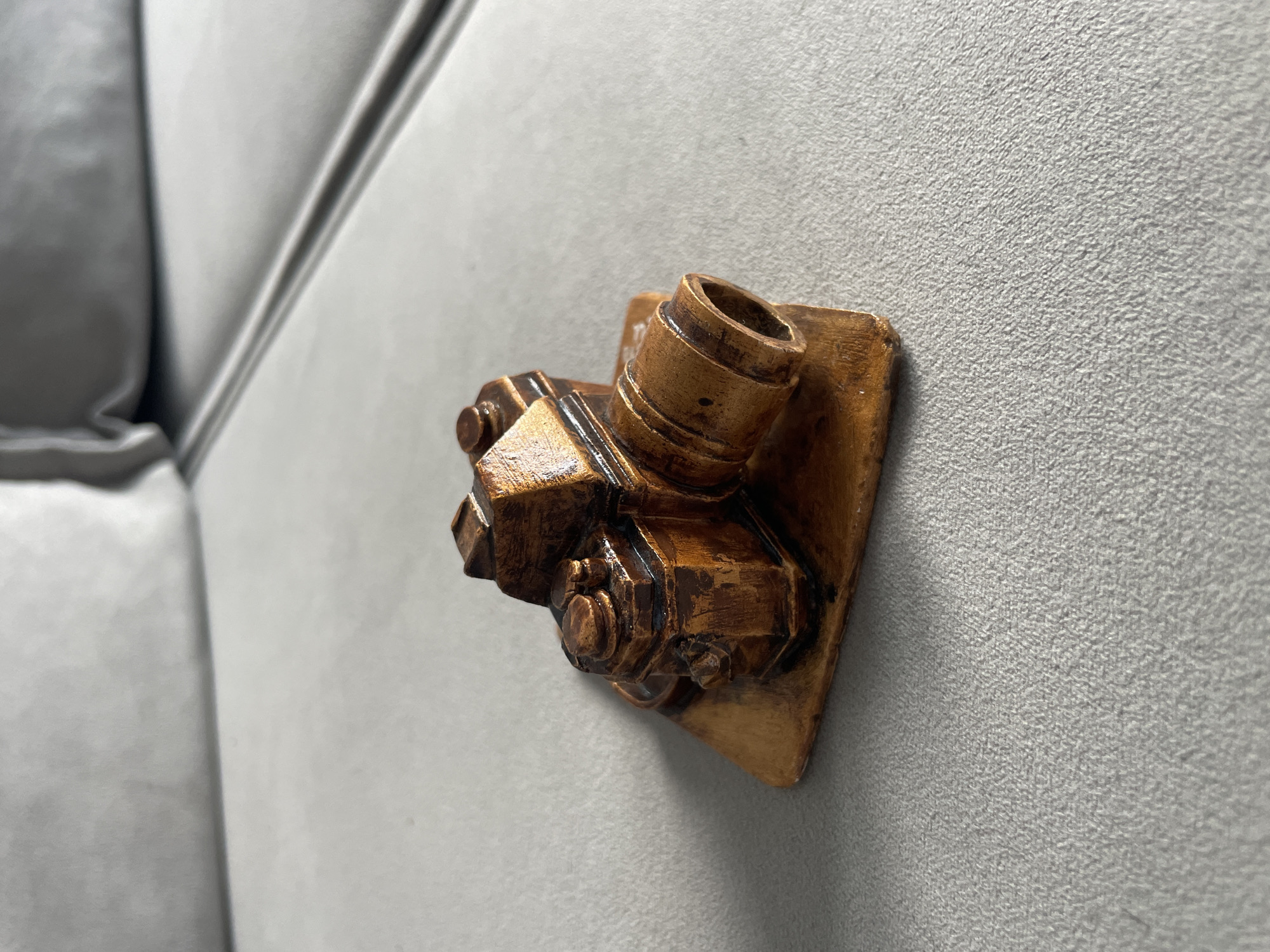 A small, wooden figurine of a camera is placed on a grey upholstered surface, likely a couch. The camera is brown and detailed, with a lens, viewfinder, and dials. The surface has a textured appearance and a slight sheen. The background is blurred, suggesting it is out of focus.