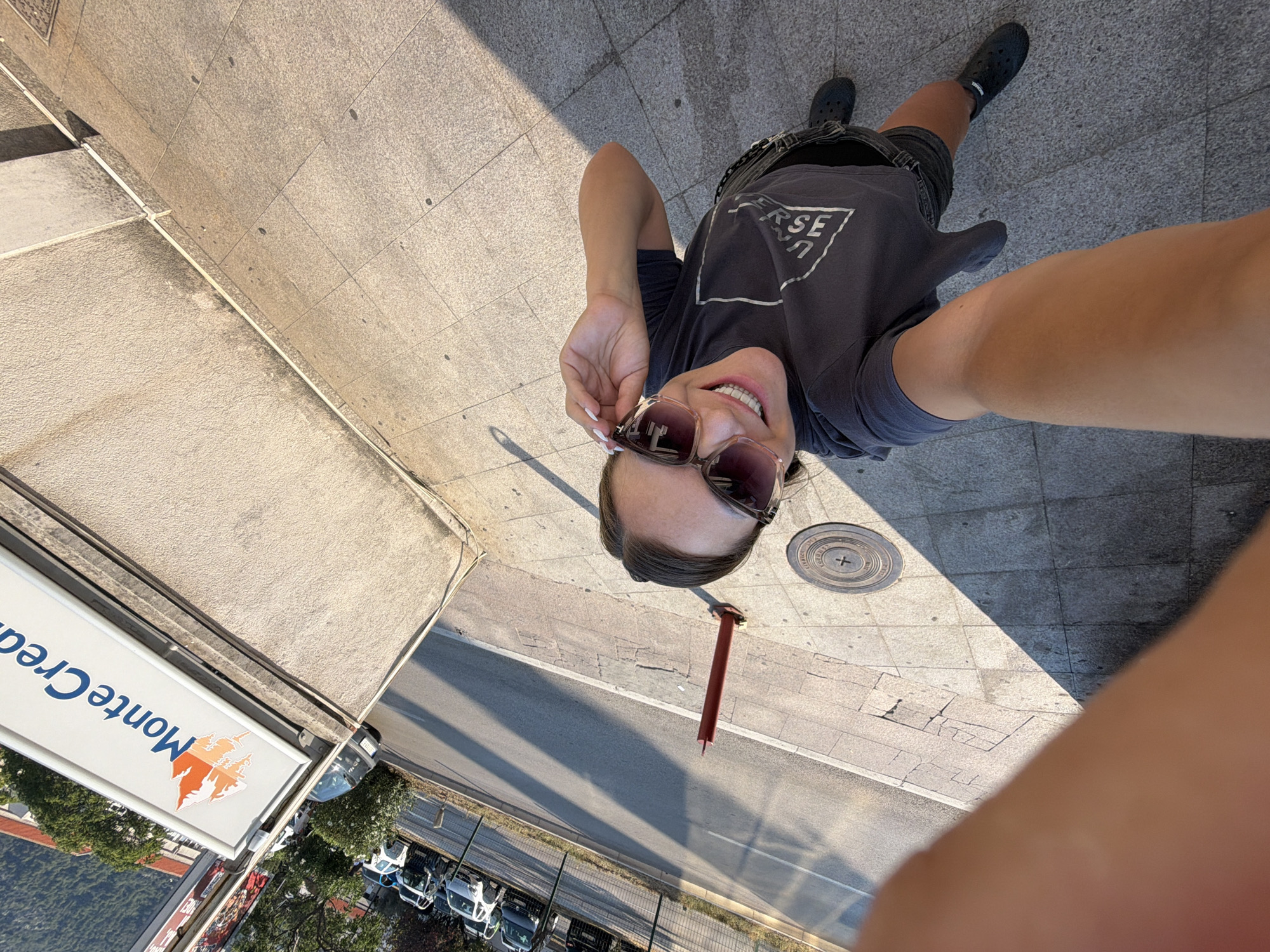 A woman is taking a selfie outdoors in Budva, Montenegro. She is wearing sunglasses, a dark t-shirt with a graphic print, and dark shorts. The background shows a sidewalk with a concrete wall, a street with parked cars, and a sign that reads "Monte Credi". The sun is casting shadows on the ground.