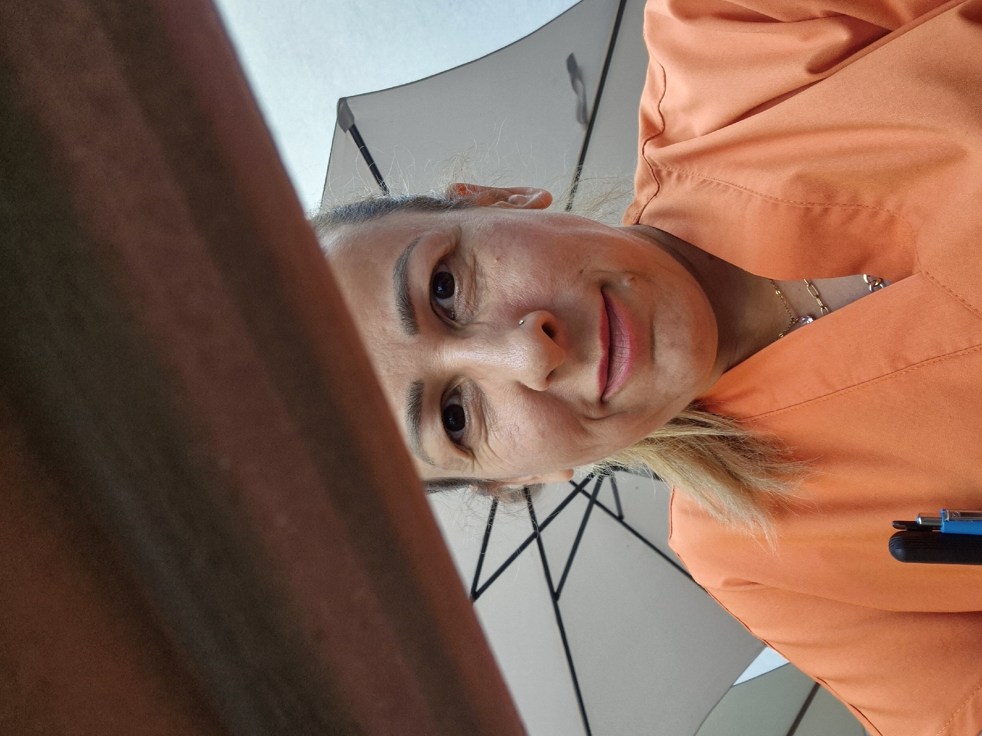 A woman, possibly in her late 30s or 40s, is photographed from a low angle, looking up at the camera. She has blonde hair pulled back, a nose ring, and is wearing an orange scrub top. A portion of what appears to be an umbrella is visible behind her, along with a dark, angled object in the foreground that partially obstructs the view of her face. She is smiling, and there is a faint sheen of sweat on her forehead and cheeks. The lighting suggests an outdoor setting, possibly during the daytime. The background is out of focus but appears to be a bright sky with some structural elements from the umbrella.