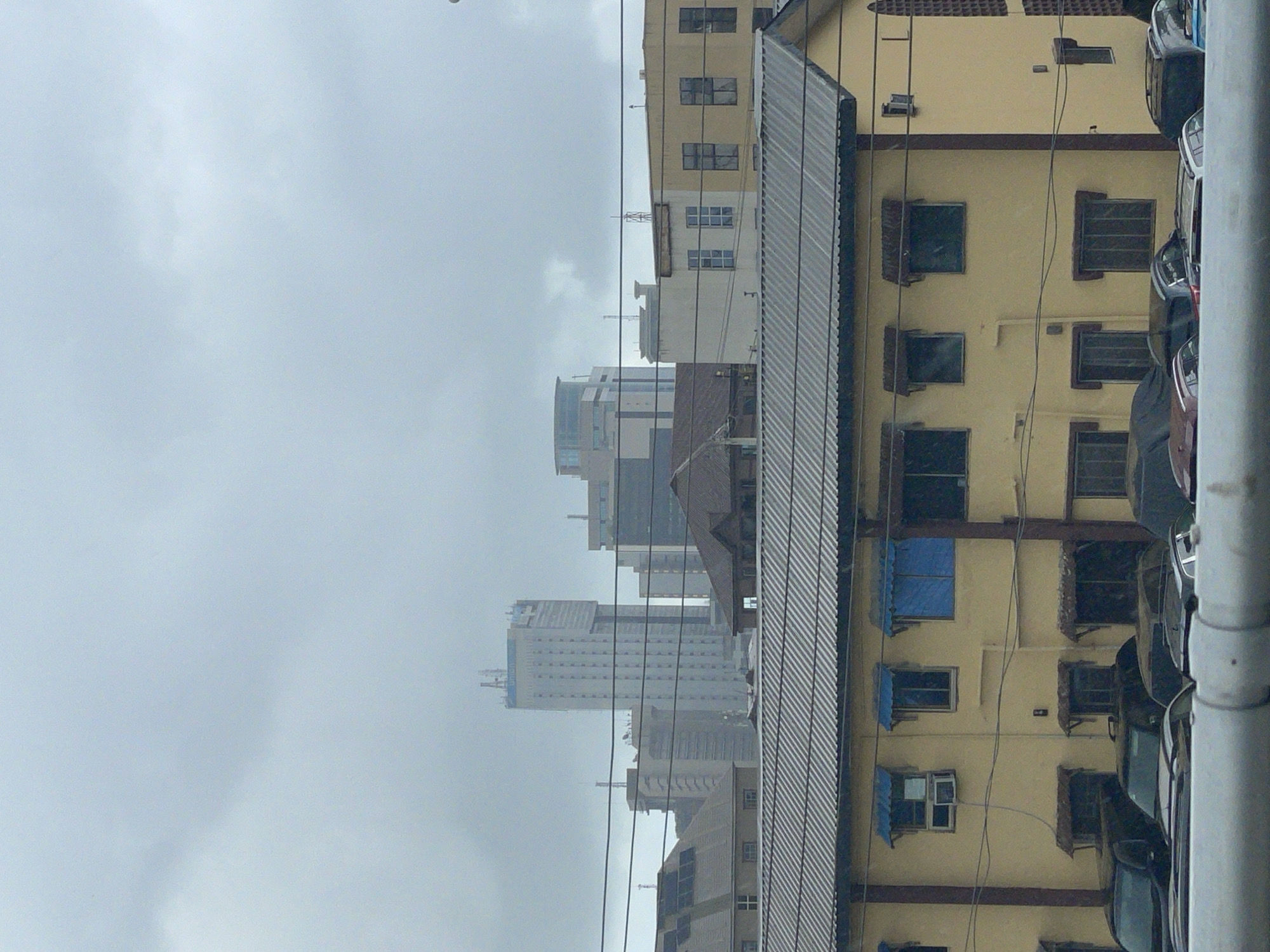 The image shows a cityscape in Lagos, Nigeria on an overcast day. In the distance are tall, modern buildings and skyscrapers, partially obscured by haze. Closer to the camera is a mid-rise yellow building with barred windows, some covered with blue tarps. Next to it is a corrugated metal structure, likely a roof. Parked along the side of the yellow building are several cars, closely packed together. Power lines crisscross the view. A thick, cylindrical metal post is visible in the immediate foreground on the right, partially obscuring the view.