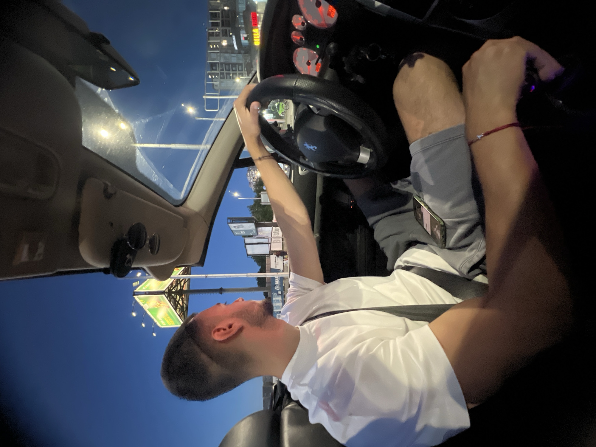 A young man with a fade haircut and a beard is driving a convertible car at dusk on a street in Plovdiv, Bulgaria. He is wearing a white t-shirt and grey shorts, with a seatbelt fastened. The car's dashboard, including the steering wheel and illuminated gauges, is visible. The surrounding environment shows streetlights and billboards, suggesting an urban setting. A phone is placed on the passenger seat. The sky is a deep blue, indicating the transition from day to night. The car appears to be moving, as indicated by the driver's focus on the road.
