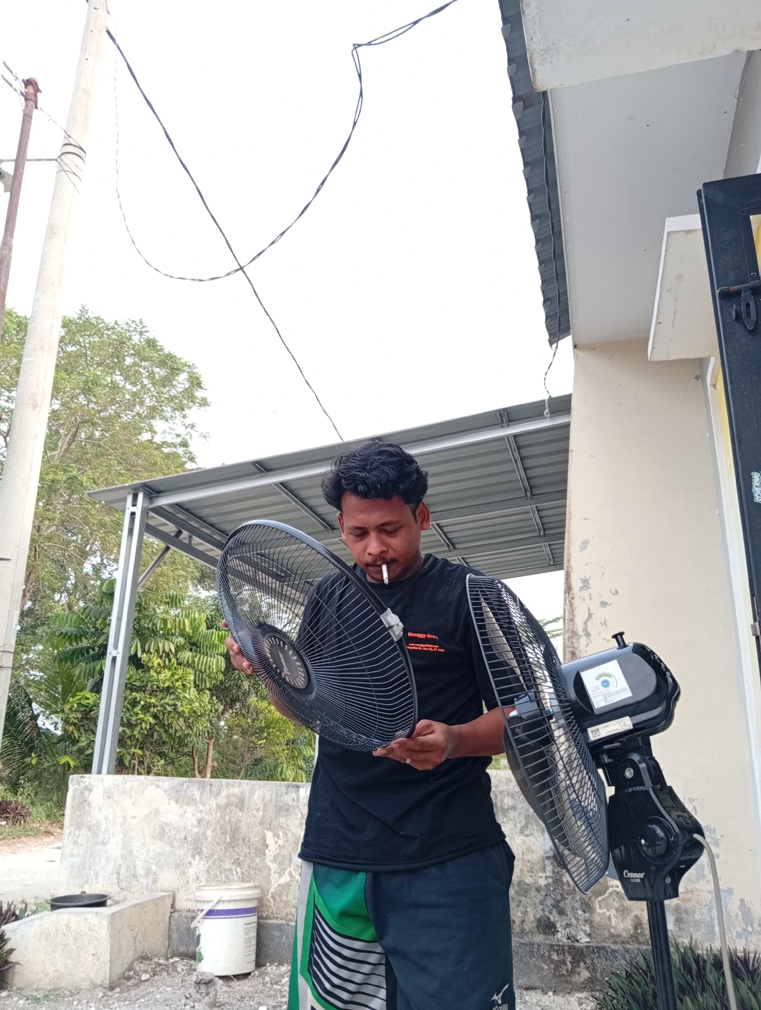 A man stands outdoors in Kualu, Indonesia, holding a fan blade in his left hand and a fan in his right. He is wearing a black t-shirt and shorts. The man is smoking a cigarette. The background shows a building, trees, and power lines. The ground is covered in gravel.