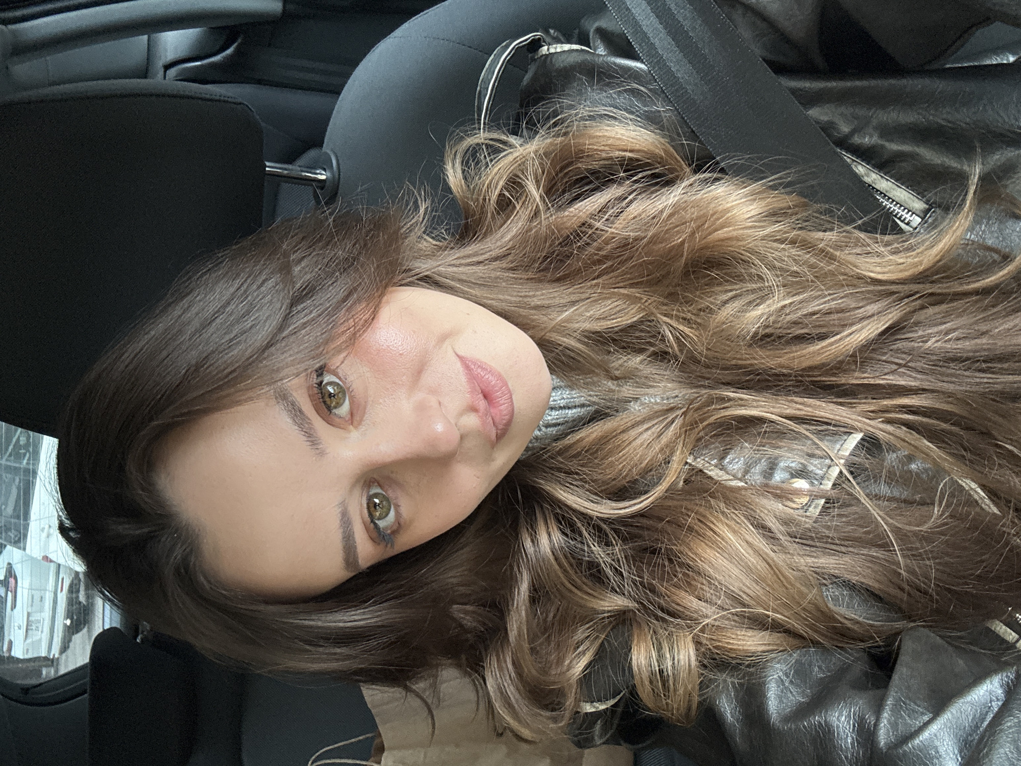 The image shows a woman with light brown hair and green eyes, wearing a black leather jacket and a grey shirt. She is wearing a seatbelt, suggesting she is inside a vehicle. The interior of the car is visible. The specific location in Krasnoyarsk, Russia, cannot be determined from the image alone.