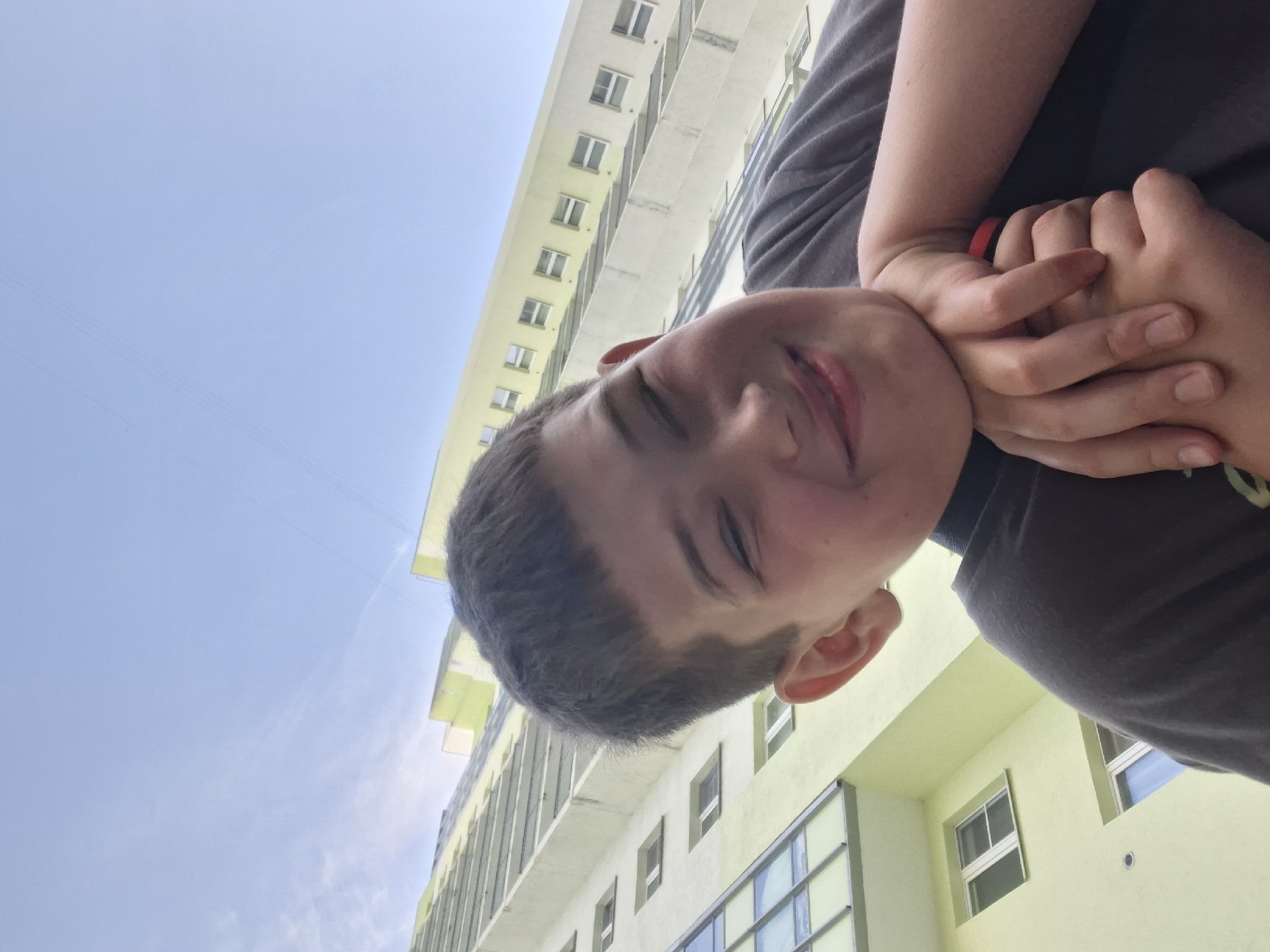 The image captures a low-angle perspective of a young boy or teenager against an urban backdrop, with the original photo appearing rotated. The boy, positioned in the foreground, looks downwards towards the camera with a playful, closed-mouth smile and partially closed eyes, conveying a happy or content emotion. He has short, dark hair and is wearing a dark-colored short-sleeved top. His hands are clasped together near his chin, adding to the relaxed posture.
Behind the boy, the frame is dominated by a bright, clear blue sky with minimal cloud cover, indicating a daytime setting. Urban architecture fills the mid-ground and background. A prominent, multi-story building with light-colored (possibly off-white or very light yellow) facades and numerous rectangular windows and balconies rises on the left side of the frame. Another building, appearing to be a light green or pale yellow, is partially visible on the right, also featuring windows. The overall impression is a cheerful moment in an urban environment under clear skies. Given the provided context, the setting is Saint Petersburg, Russia. There is no visible text in the image.