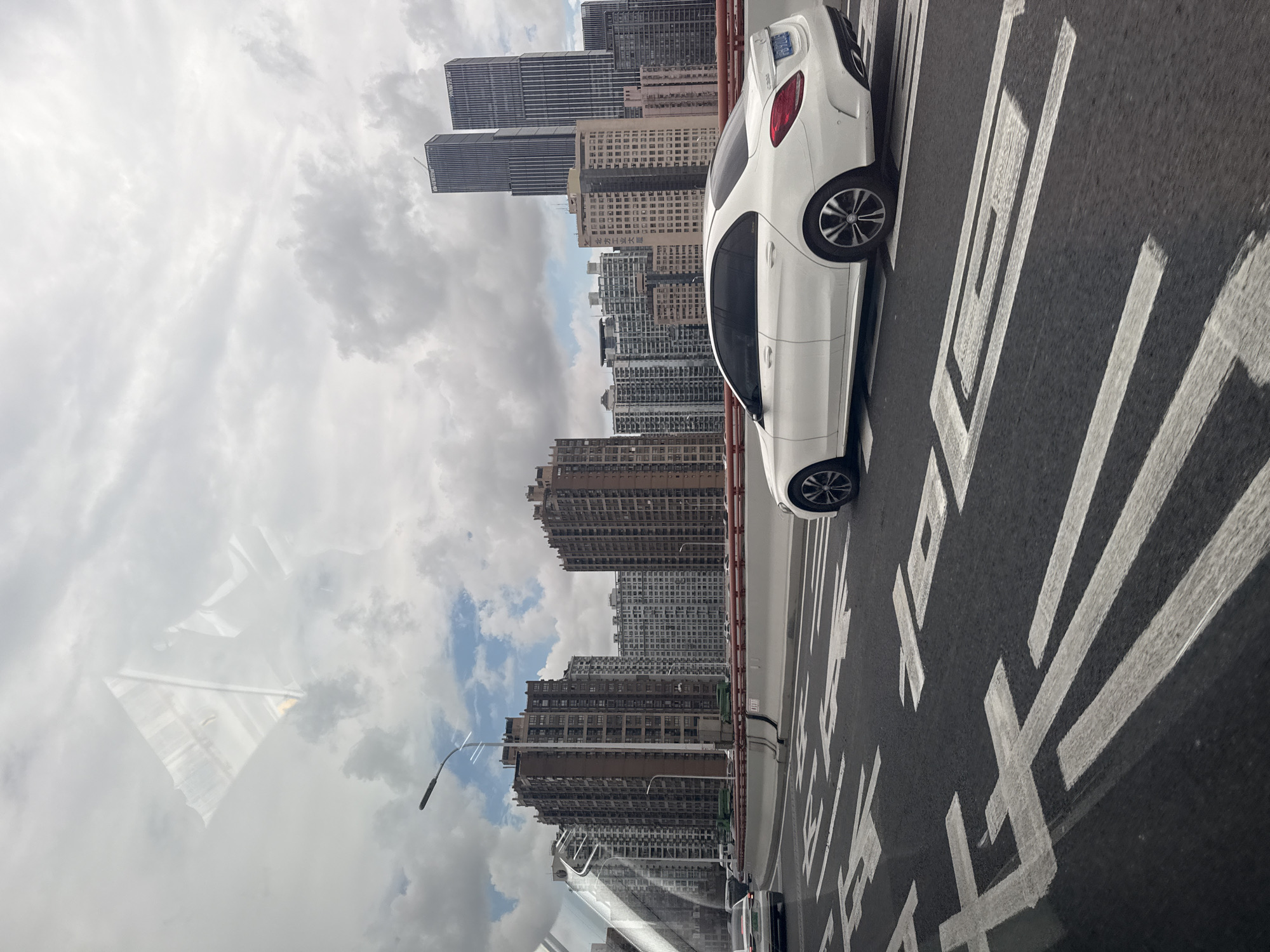 This image depicts a view from a moving vehicle on a bridge in Shanghai, China, likely the Nanpu Bridge. In the foreground, the asphalt road displays white lane markings, including what appear to be directional arrows. A white Mercedes-Benz C-Class sedan is the primary subject in the foreground, traveling in the middle lane. The background features a dense urban skyline characterized by numerous high-rise buildings of varying architectural styles and heights. Two prominent modern skyscrapers with vertical striped facades stand out in the mid-distance. The sky is overcast with scattered clouds, suggesting a moderate weather day. The scene is dynamic, capturing a snapshot of urban transportation and the city's built environment.
