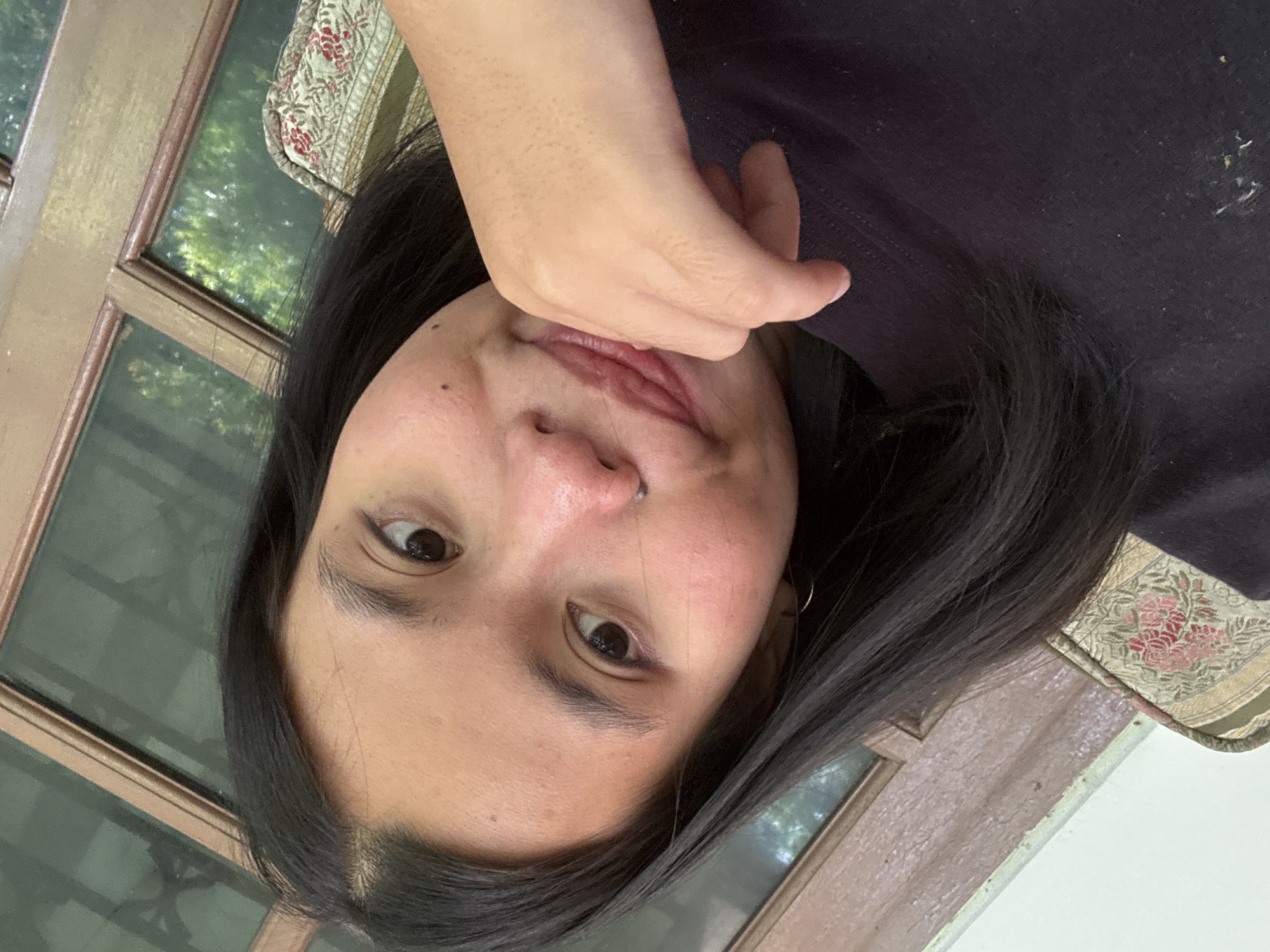 A young woman with dark hair and light brown skin is captured in a close-up, slightly angled selfie. She is wearing a dark t-shirt and has her right hand resting on her chin, her thumb touching her lips. A small silver hoop earring is visible in her left ear, and she has a small mole above her lip. The background shows a wooden window frame and greenery, suggesting an outdoor or semi-outdoor setting with foliage visible through the glass. The image appears to be taken in a residential area, potentially a home or a cafe, in South Jakarta, Indonesia, given the context. The lighting is soft and natural, highlighting the woman's features and the textures of her surroundings.