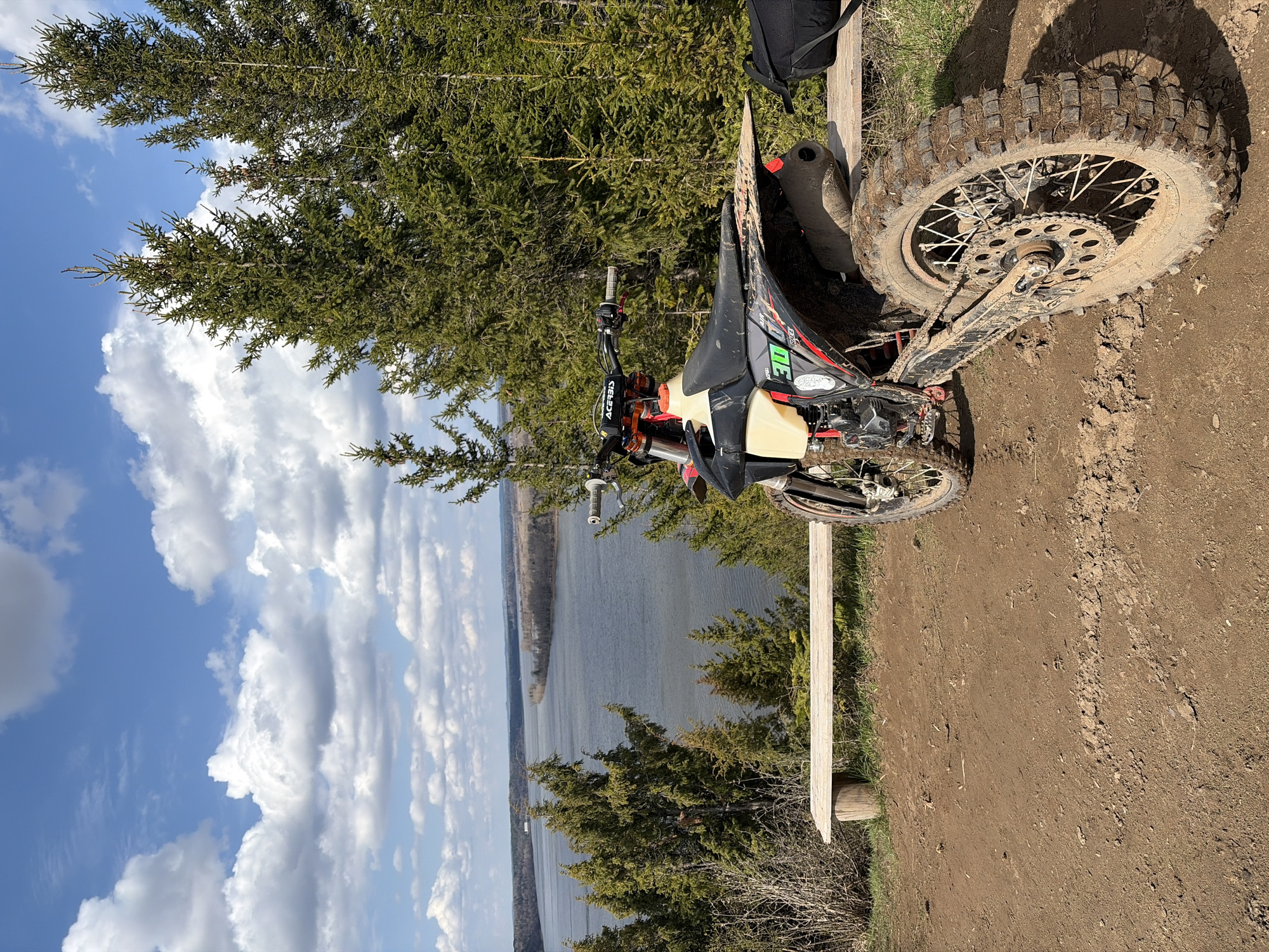 The image depicts a muddy dirt bike parked on a dirt track overlooking a body of water. Lush green trees frame the scene. The sky is blue with scattered clouds. A wooden bench sits near the dirt bike. The location may be in or near Ольховка, Russia.