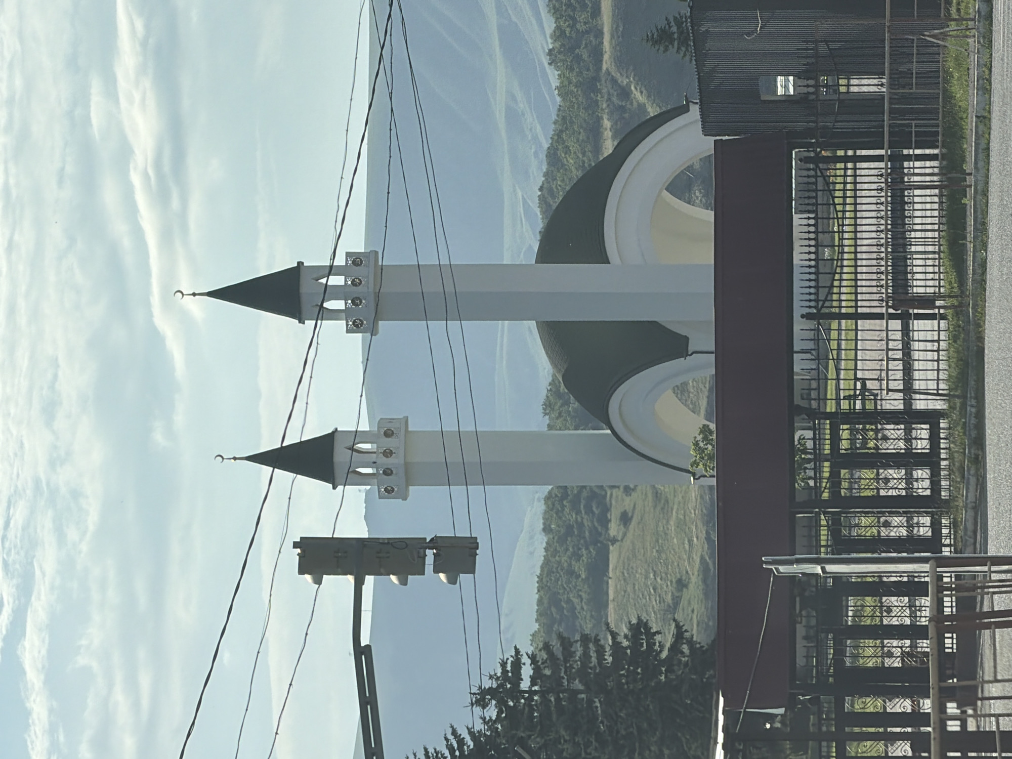 This vertical image captures an outdoor scene in Kubina, Russia, featuring a mosque with two prominent minarets against a backdrop of rolling hills and a sky filled with wispy clouds. The mosque's minarets are white and slender, topped with dark, conical roofs adorned with crescent moons. A large, dark green dome is partially visible between the minarets. In the foreground, to the left, a traffic light is mounted on a pole. To the right, a black metal fence with ornate scrollwork encloses an area, and beyond that, a corrugated metal structure with a small window is visible. The scene appears to be set during the daytime, with bright, diffused light suggesting it could be morning or late afternoon. The weather seems clear, with the sky predominantly blue with white clouds. The overall impression is one of tranquility and architectural beauty in a natural landscape. There is no visible text in the image.